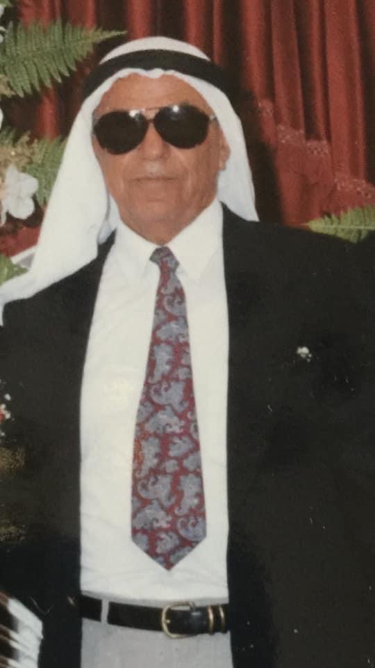 Man wearing sunglasses, white headscarf, black suit, white shirt, and patterned tie, standing indoors with red curtains and plants behind.