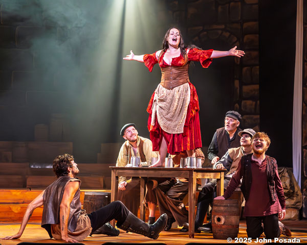 Woman in costume stands on table with arms outstretched, surrounded by five people on stage during a theatrical performance.