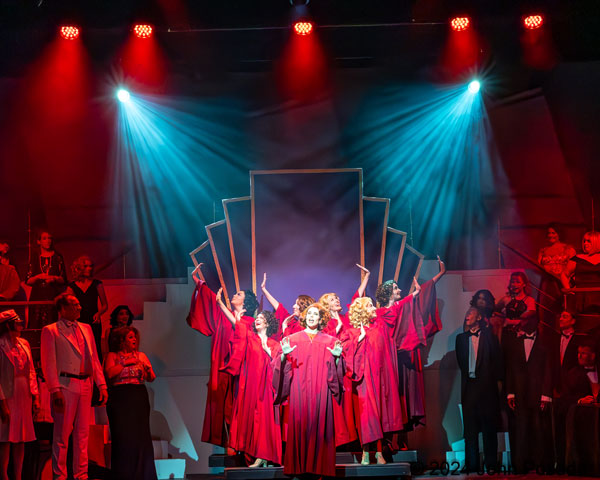 Chorus of people in red robes on stage with blue and red lighting, some holding hands, in a theatrical performance.