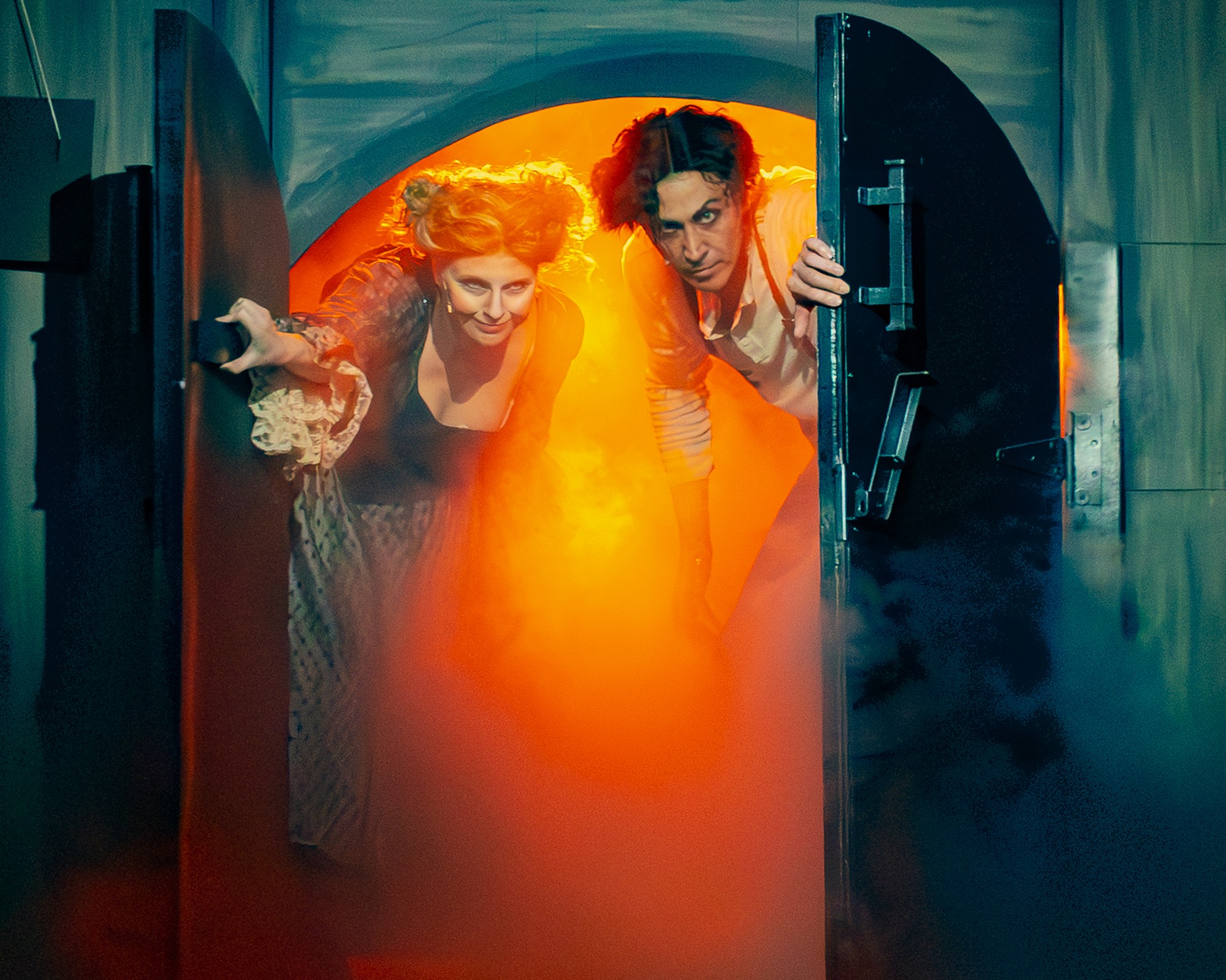 Two people look through a door with orange glowing light and smoke, smiling and leaning forward.