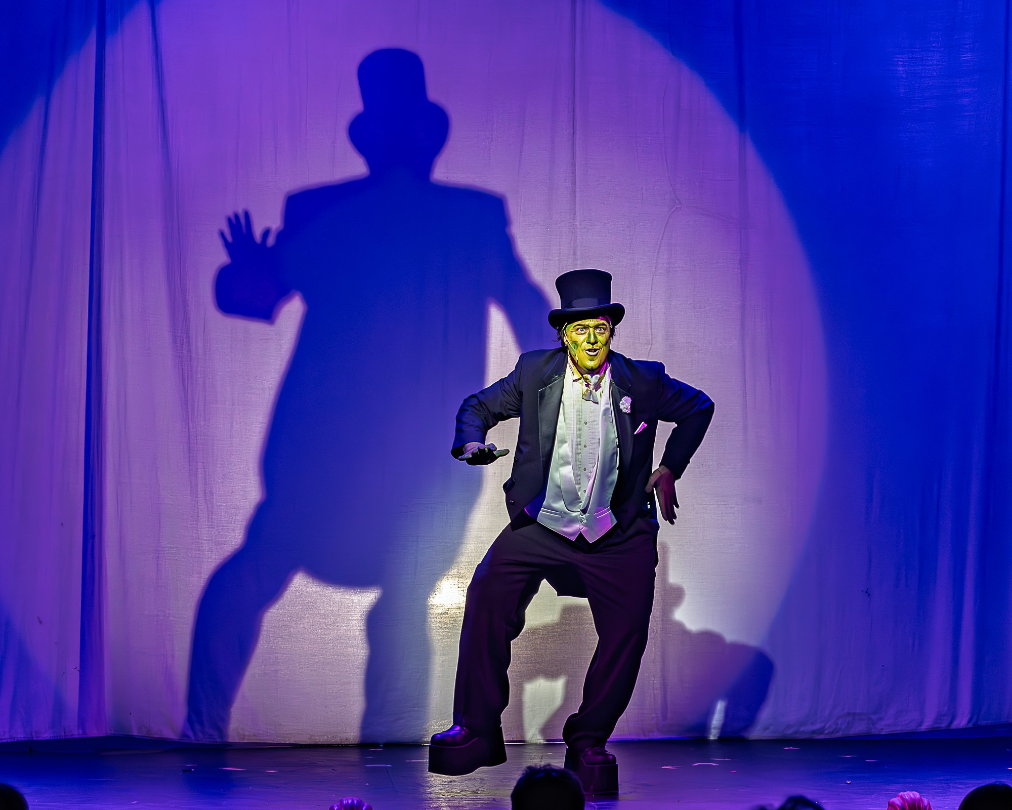 Performer in a tuxedo and top hat dances on stage with a large shadow cast on curtain behind, spotlight focused on him.