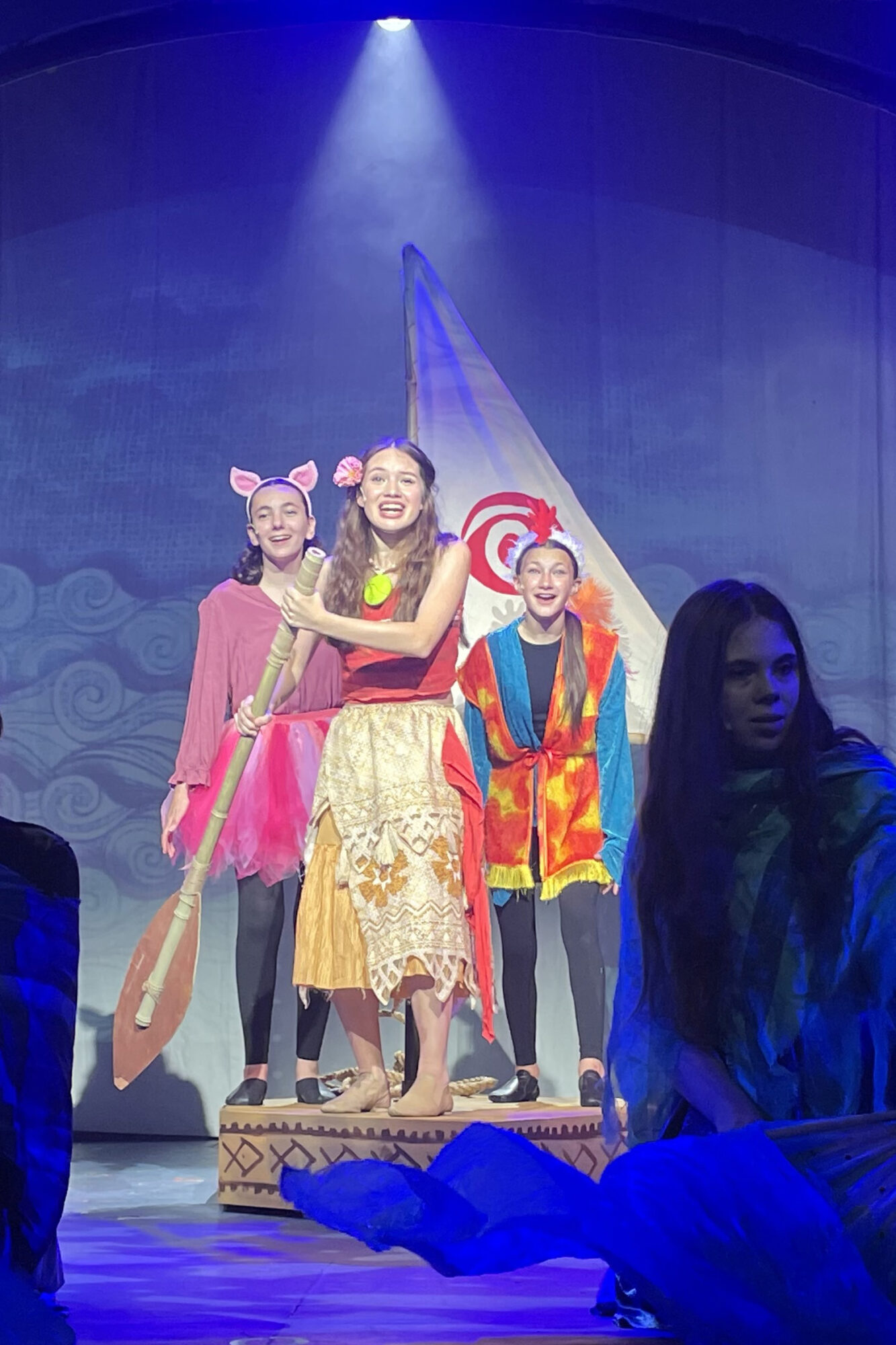 Group of children performing on stage, dressed in colorful costumes, with a blue spotlight overhead.
