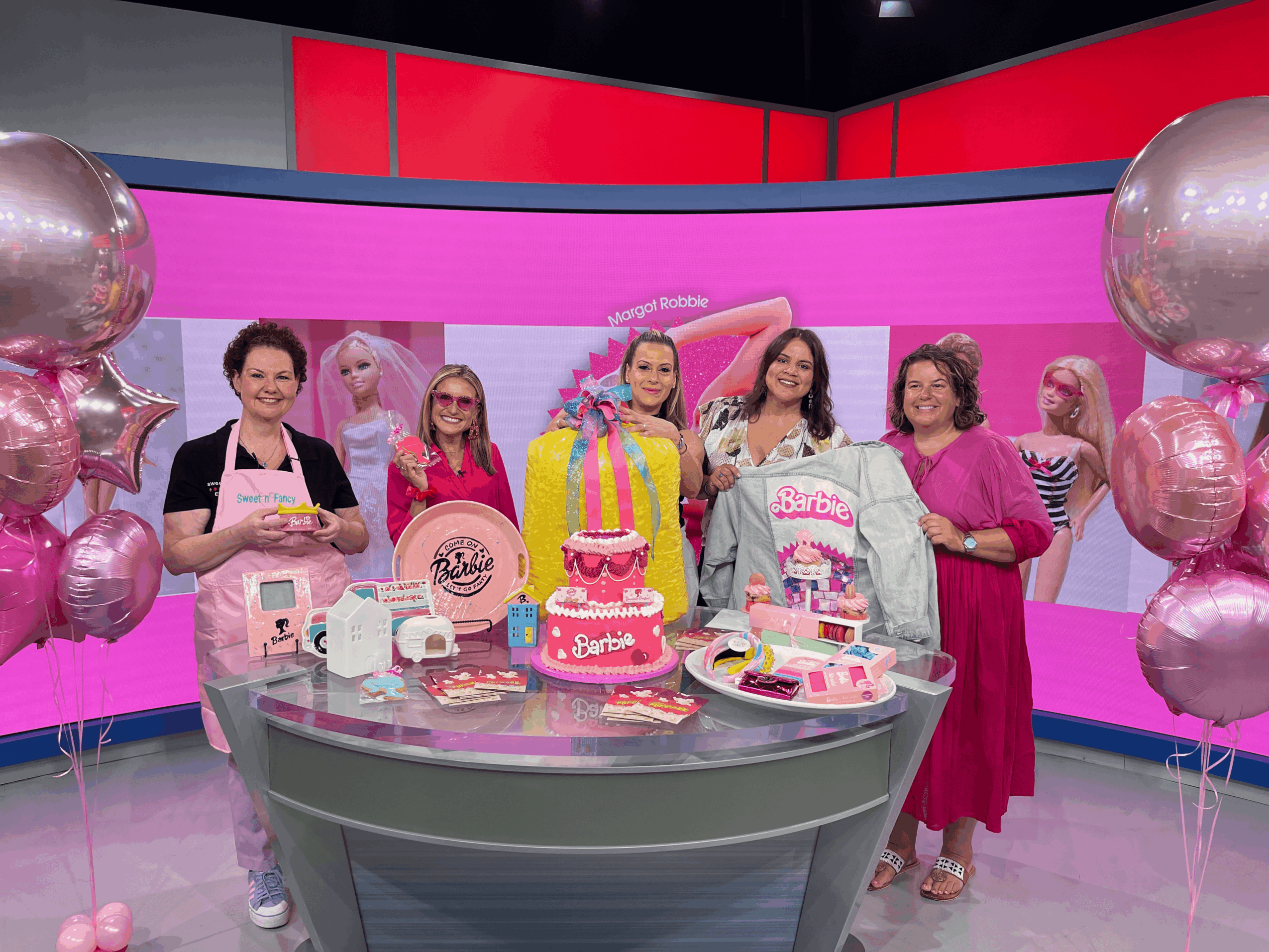 Five women stand behind a table with a pink Barbie-themed cake, toys, and decorations, in a colorful studio setting.