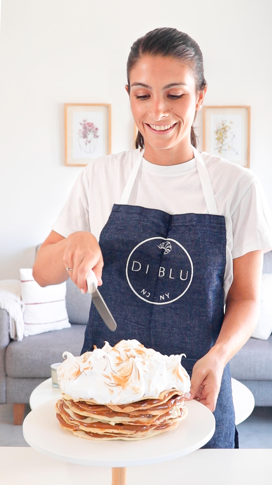 Woman smiling, wearing apron, slicing a layered cake on a white plate in a bright room.