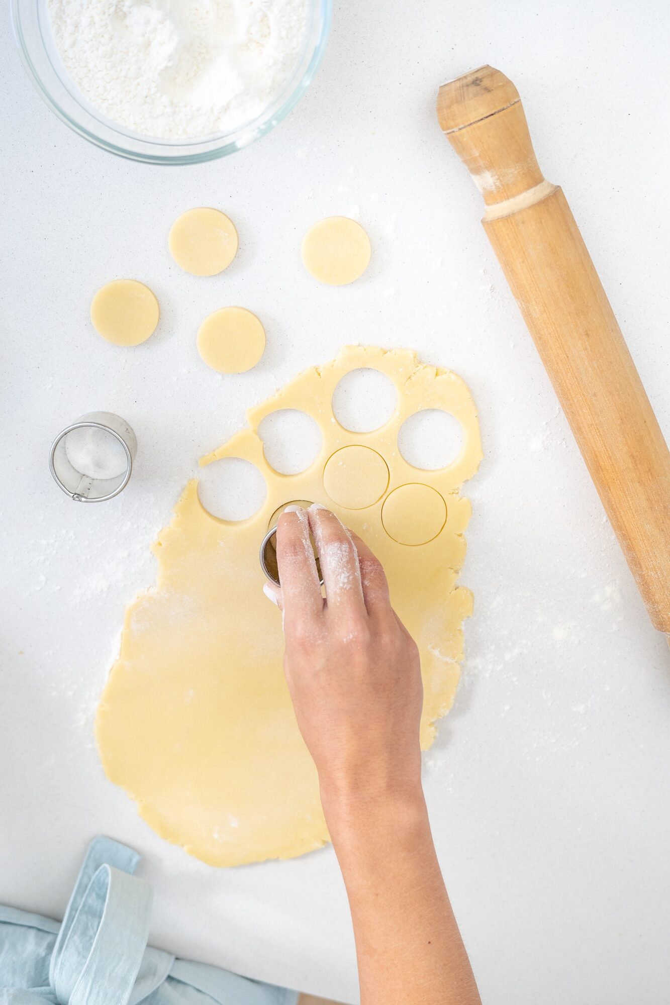 Person's hand cutting dough with a round cutter, with baking tools and ingredients on a white surface.