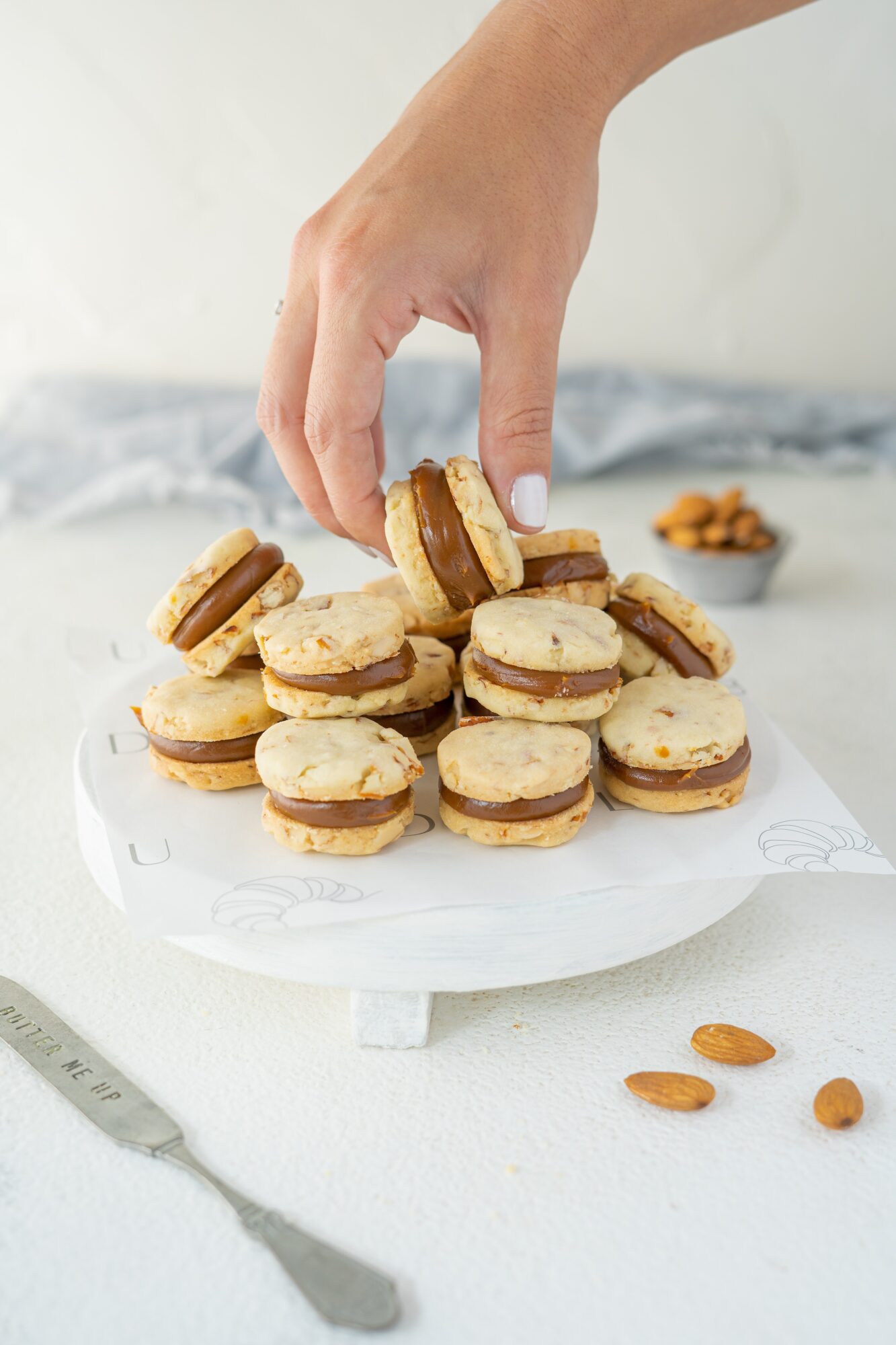 Hand reaching for stacked cookies with cream filling on a white plate, with almonds and a knife nearby.