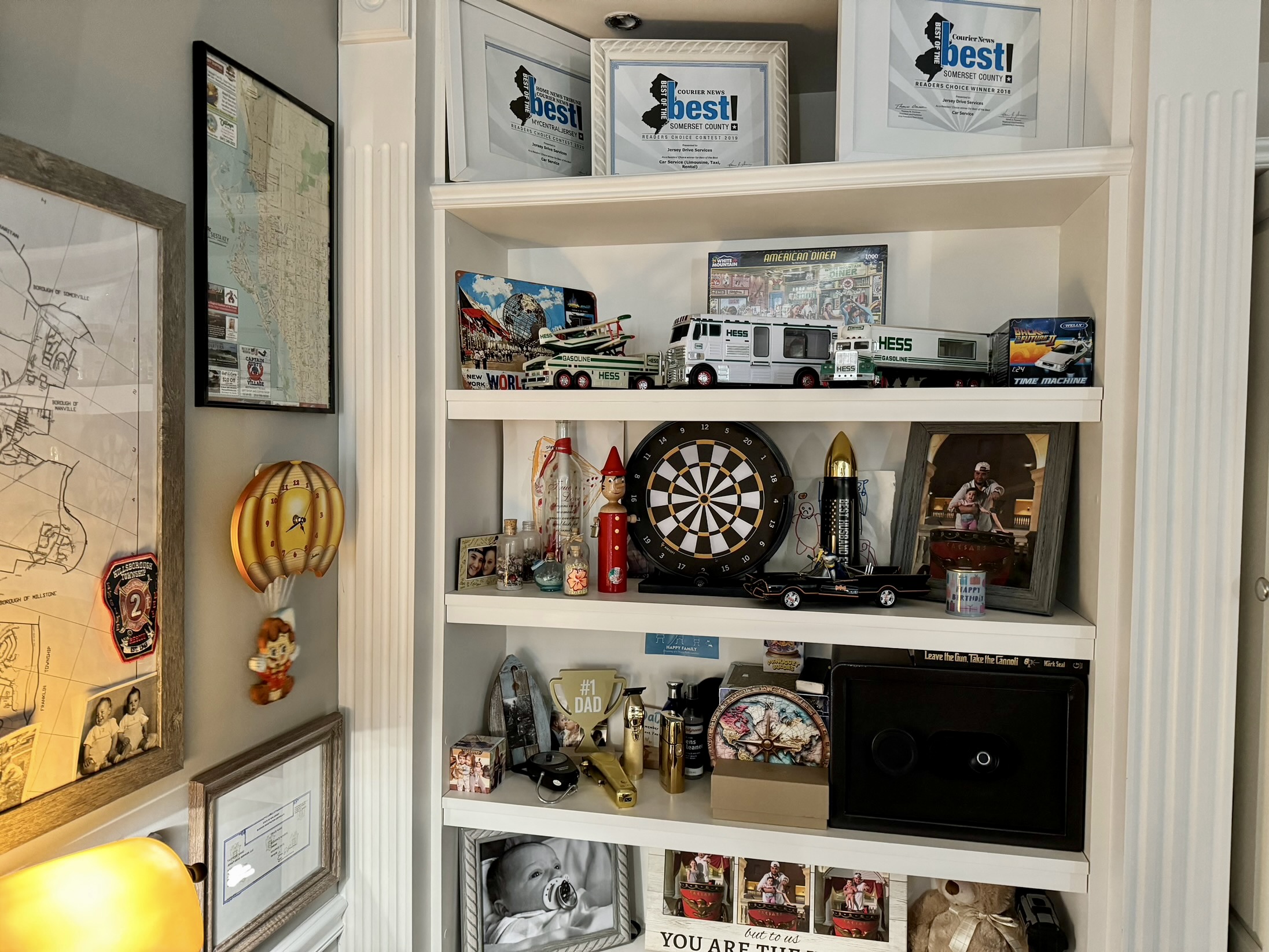 White bookshelf with framed photos, model vehicles, a dartboard, and decorative items, against a wall with a mirror and artwork.