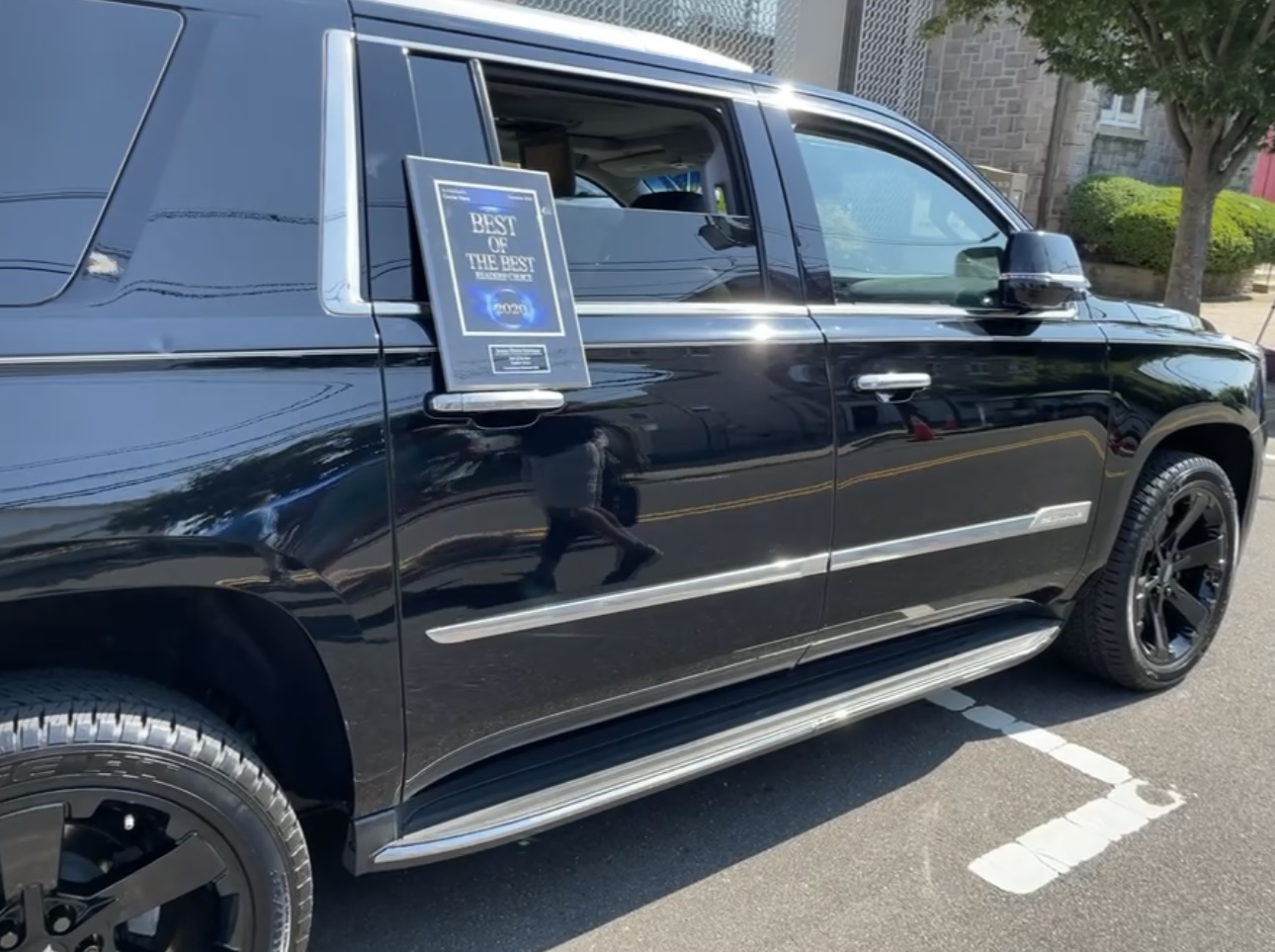 Black SUV parked in a parking lot with a sign on the window, trees and buildings in the background.