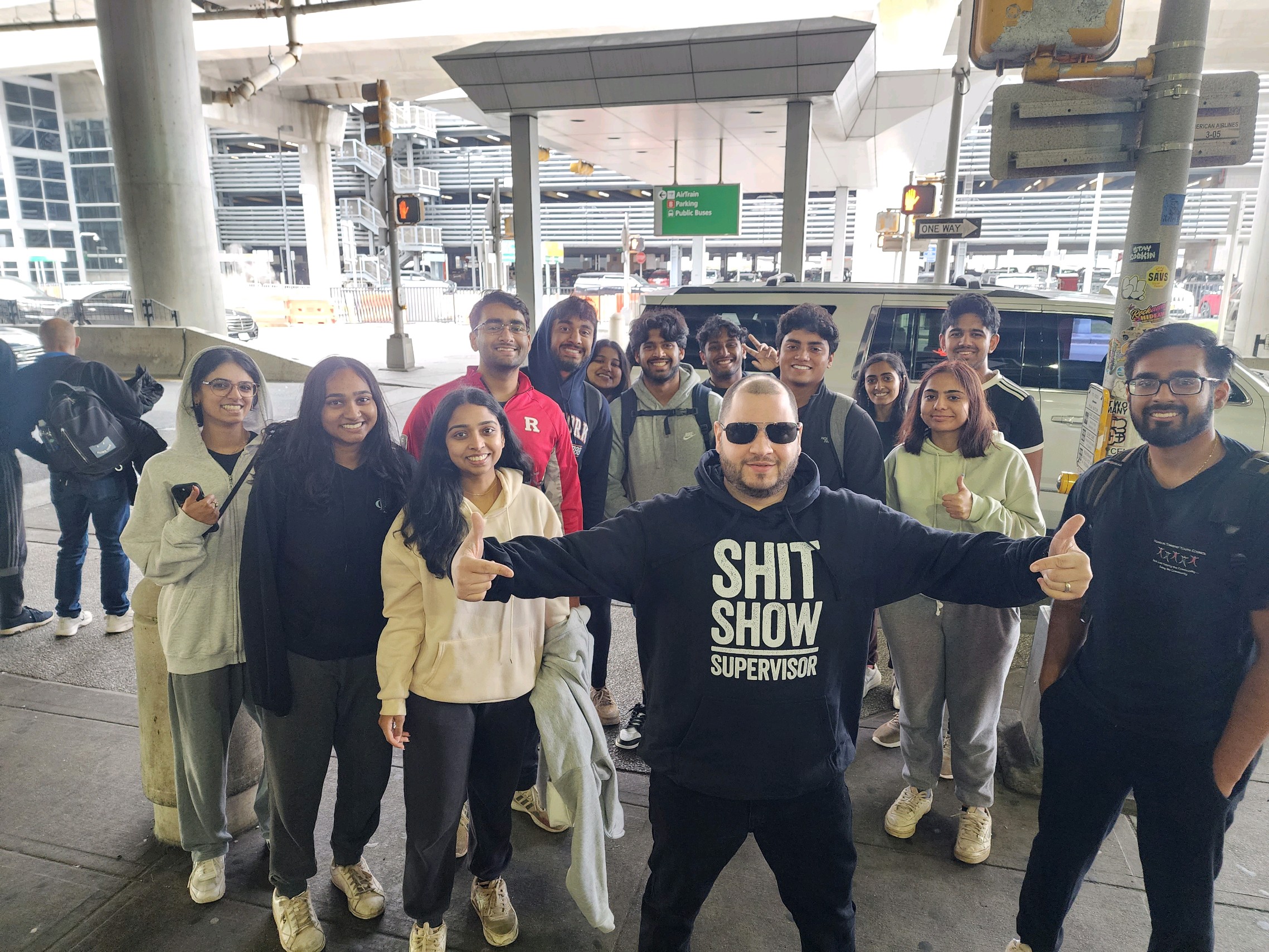 Group of people standing outdoors at an airport, smiling and posing for a photo, with a man in front wearing a black hoodie.