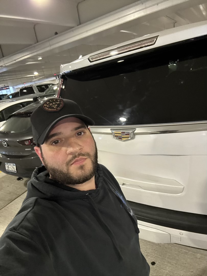 Man with a beard wearing a black cap and hoodie taking a selfie in a parking garage with a large white vehicle behind him.