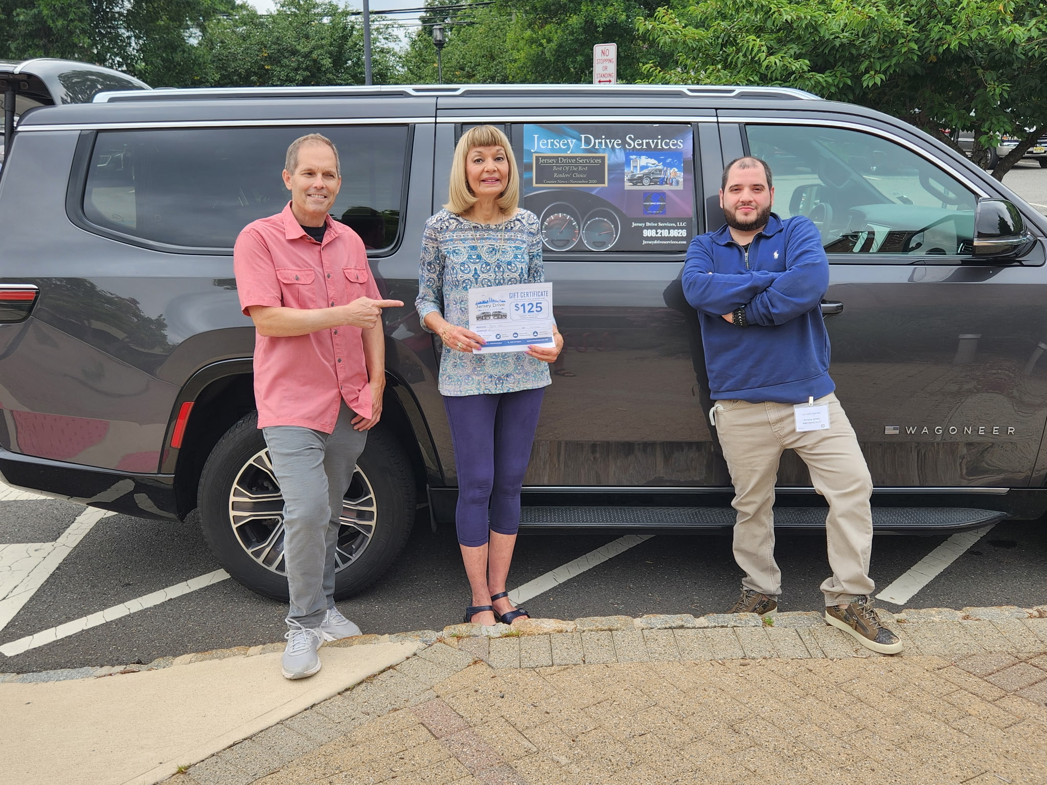 Three people stand in front of a black van outdoors, smiling, with trees and a parking lot in the background.