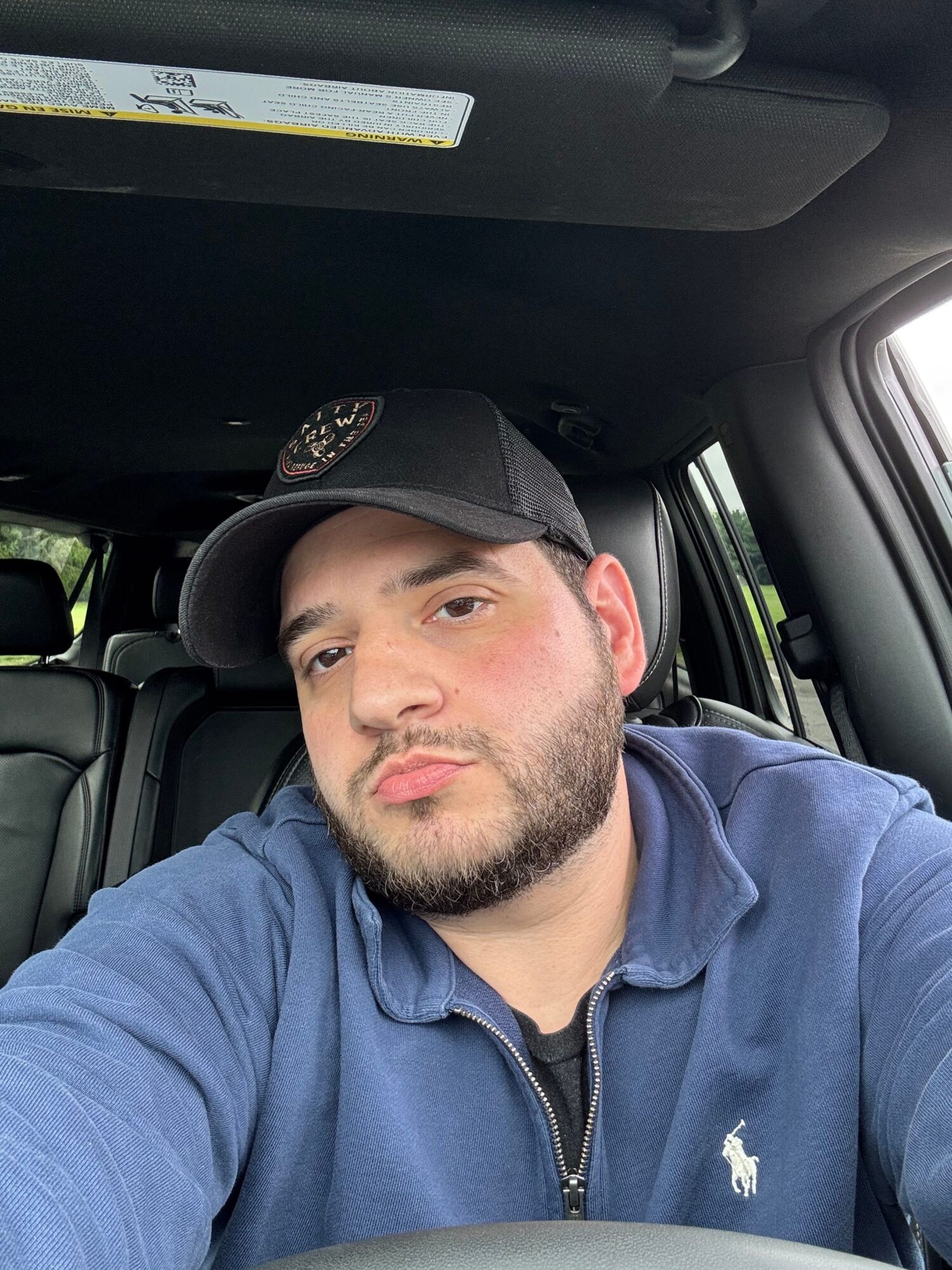 Man with beard and cap sitting in car, looking at camera, wearing a blue jacket, head resting against car seat.