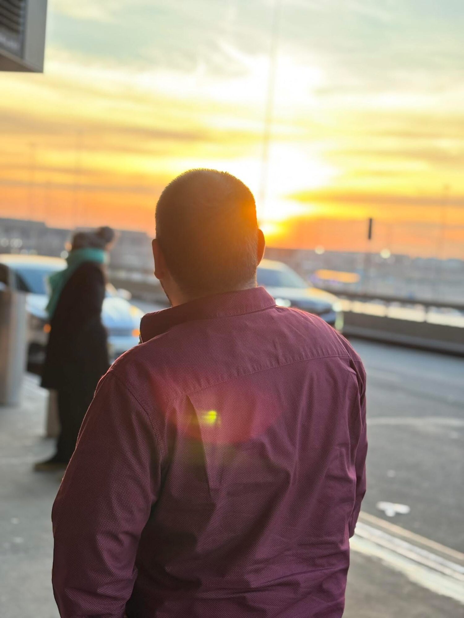 Person with short hair wearing a pink shirt standing outdoors during sunset, with cars and a person in the background.