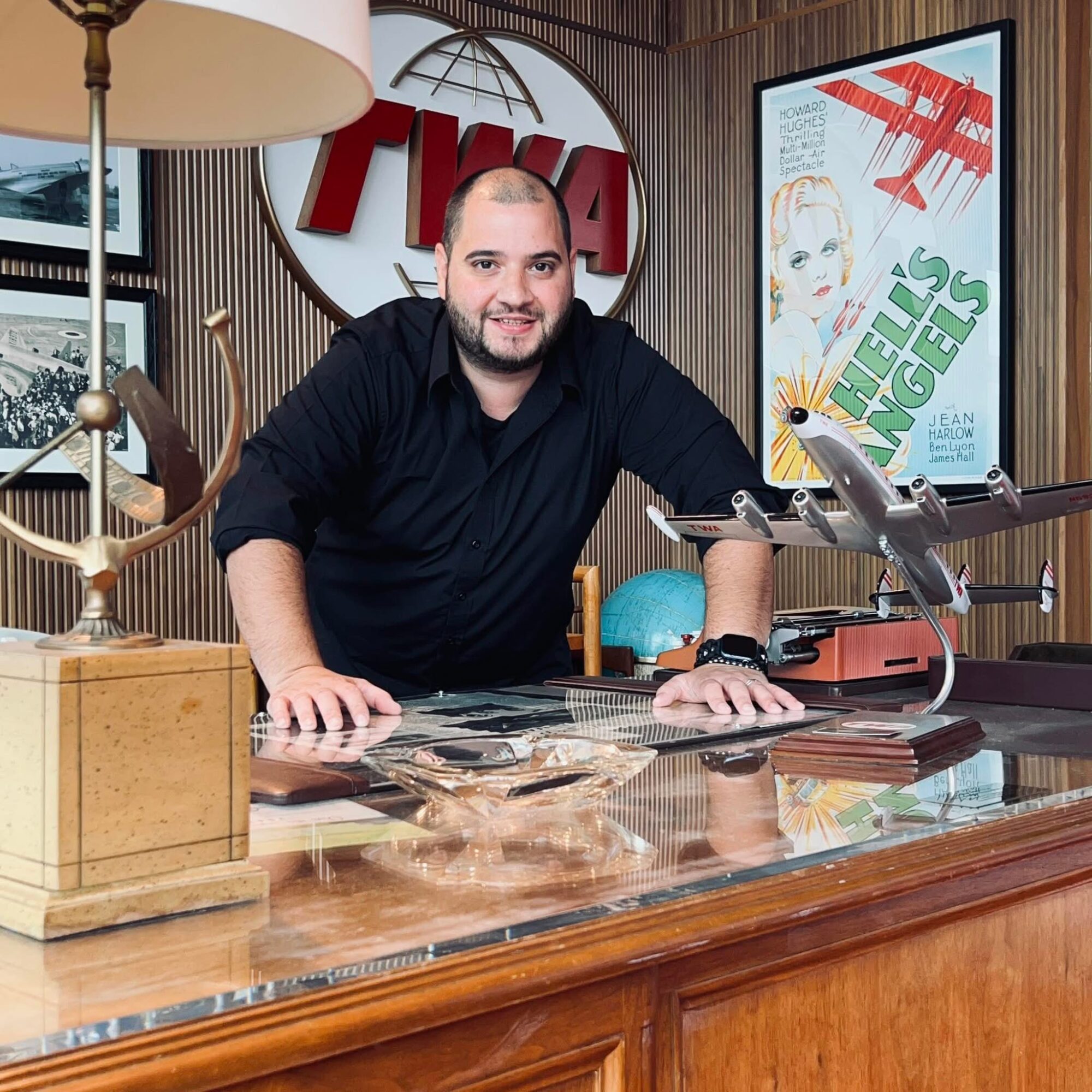 Man with beard and black shirt leaning on a wooden desk in a room with TV screens and decor.