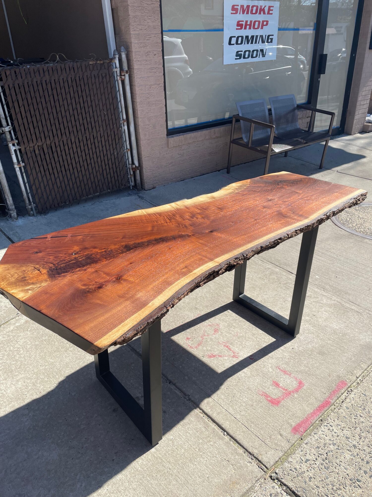 Wooden table with a natural edge, placed outdoors on a sidewalk, casting a shadow, with a storefront window nearby.