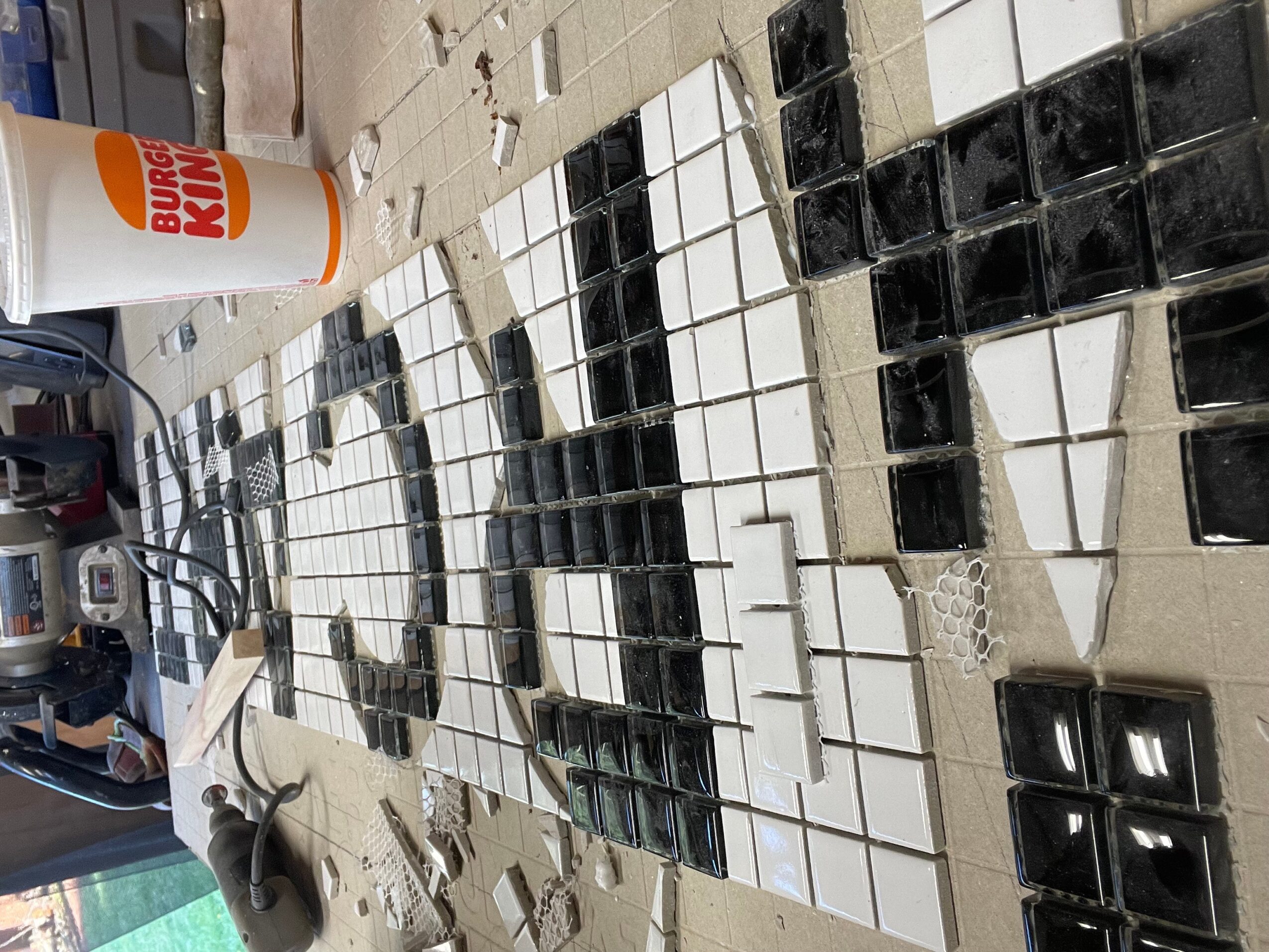 Mosaic tile artwork in progress with black, white, and gray tiles, a glue bottle, and tools on a work surface.