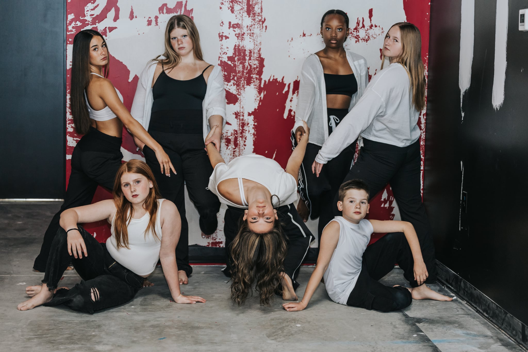 Seven young women pose against a graffiti-covered wall, some sitting and some standing, in casual clothing. Word 'LOVE' partially visible on wall.