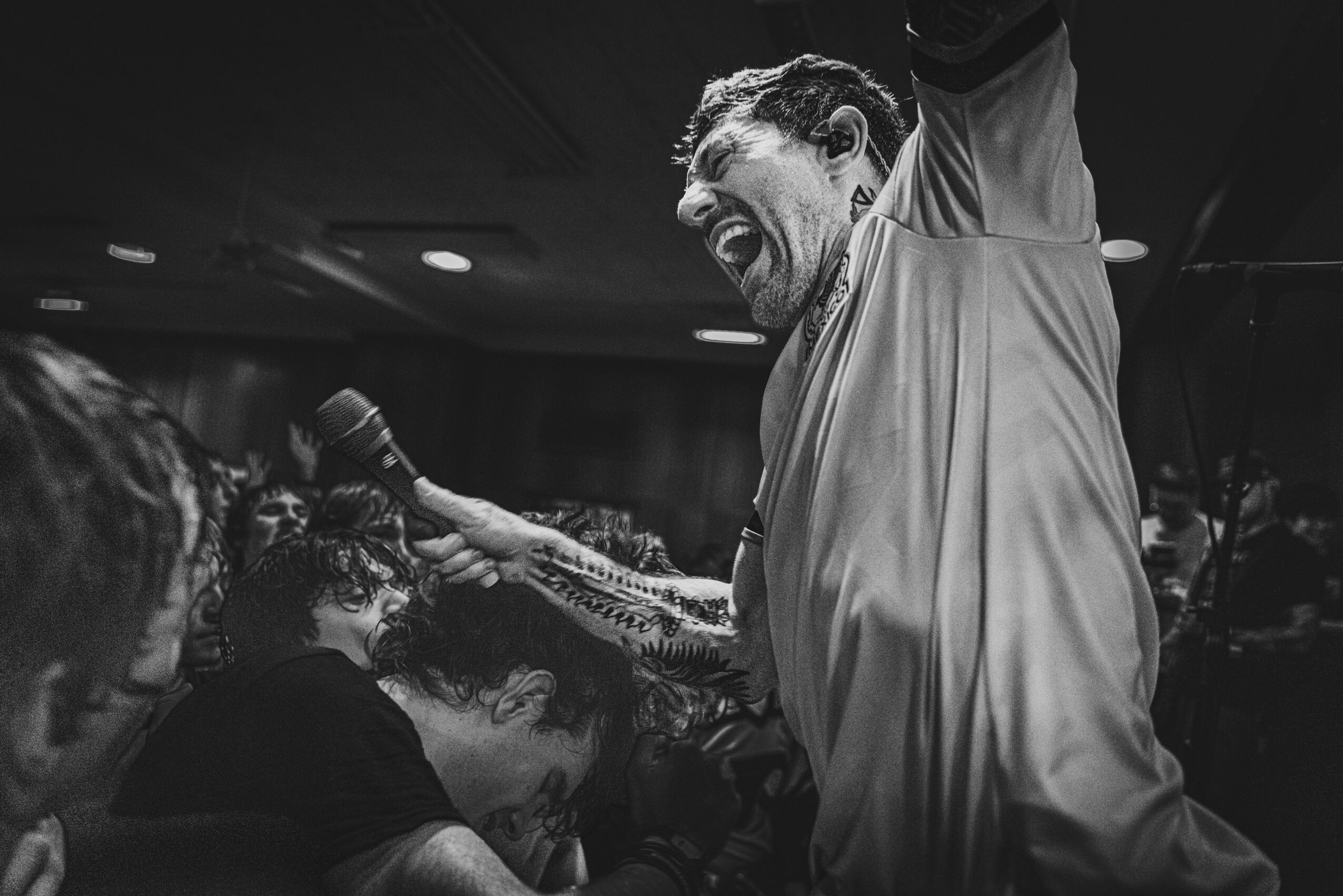 Person with long hair and beard passionately singing or shouting into a microphone at a lively event, surrounded by others.