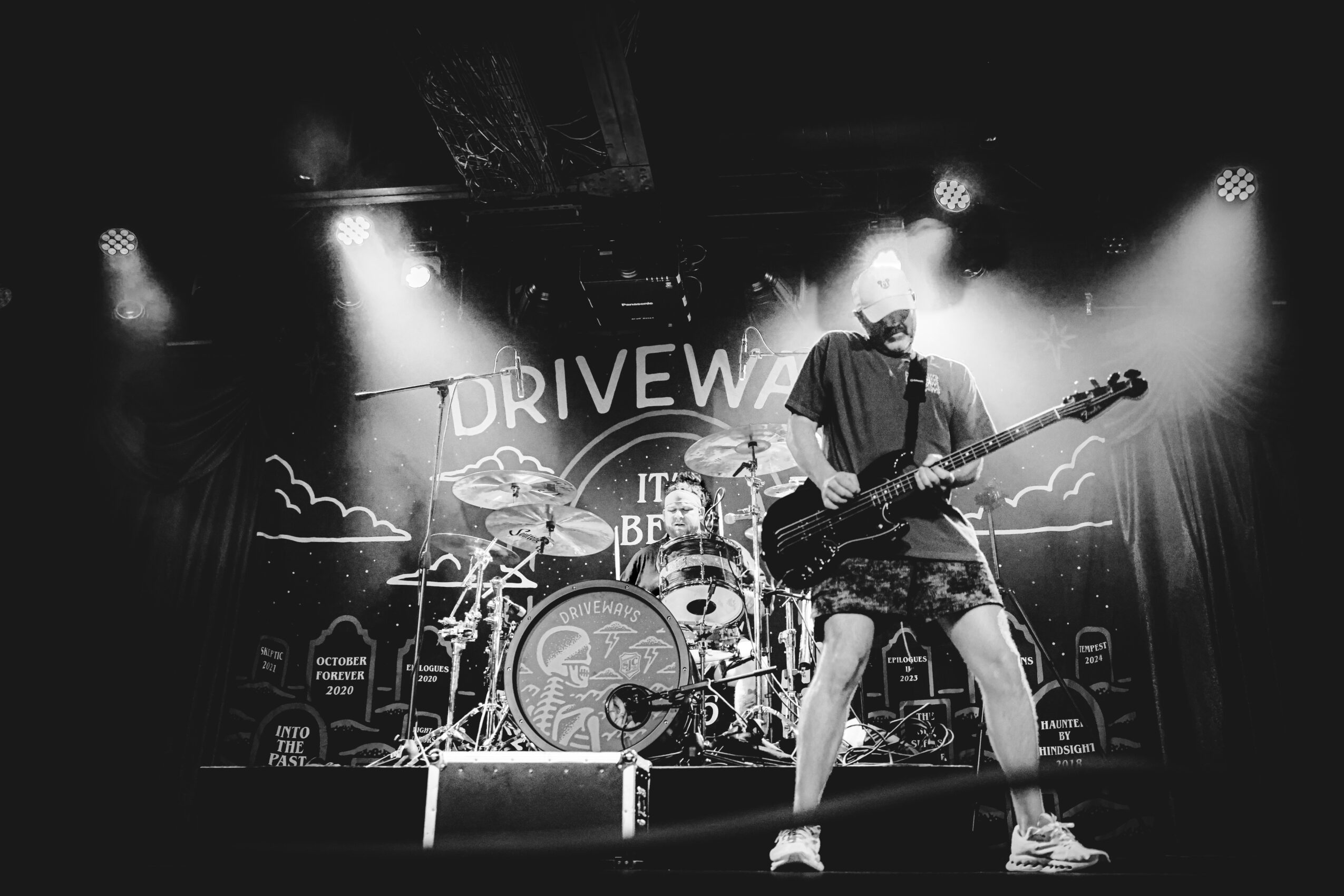 Musician playing guitar and singing on stage with drums behind, illuminated by stage lights, with a backdrop reading 'DRIVEWAY'.