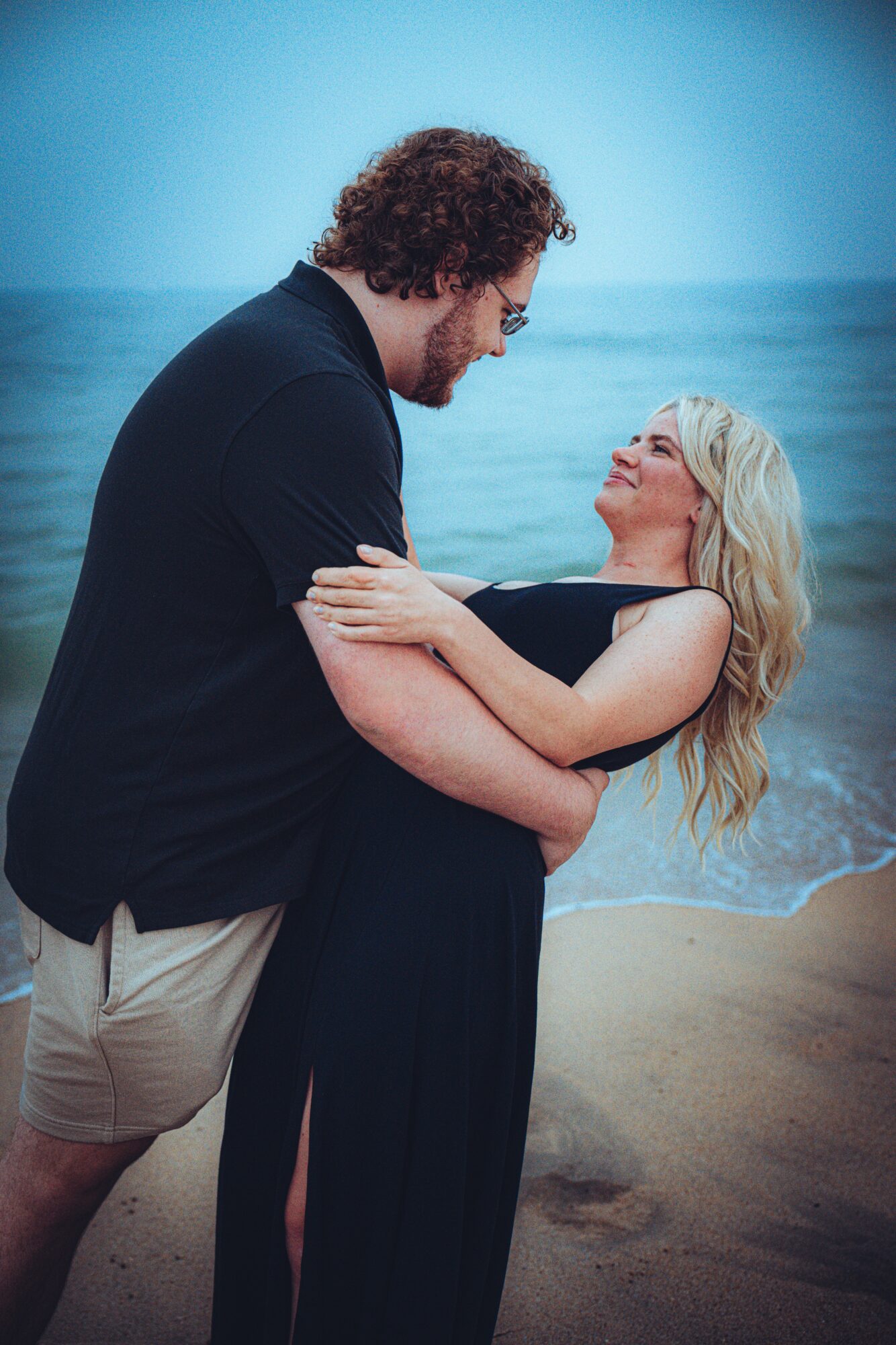 A man and woman embrace on a beach with ocean in background, smiling at each other.
