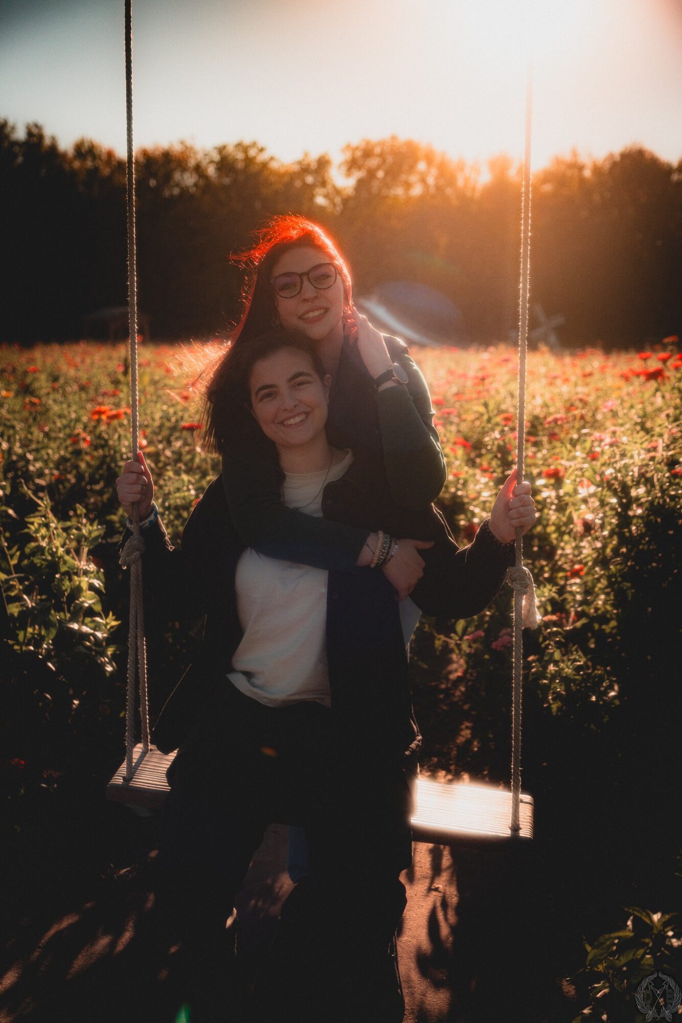 Two people sitting on a swing in a field during sunset, smiling and embracing each other.