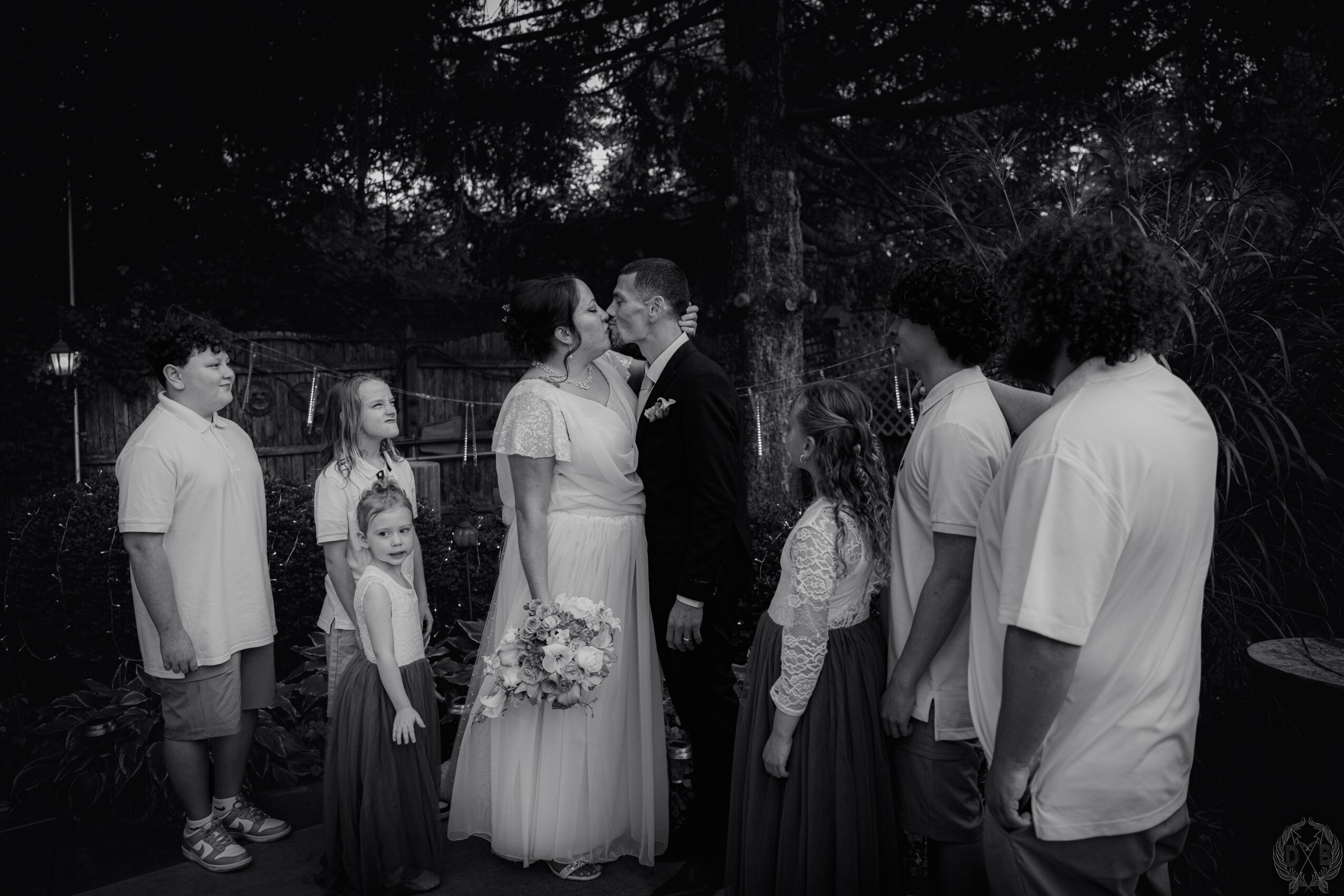 Group of people gathered outdoors, with a couple in the center, surrounded by children and adults, trees in background.