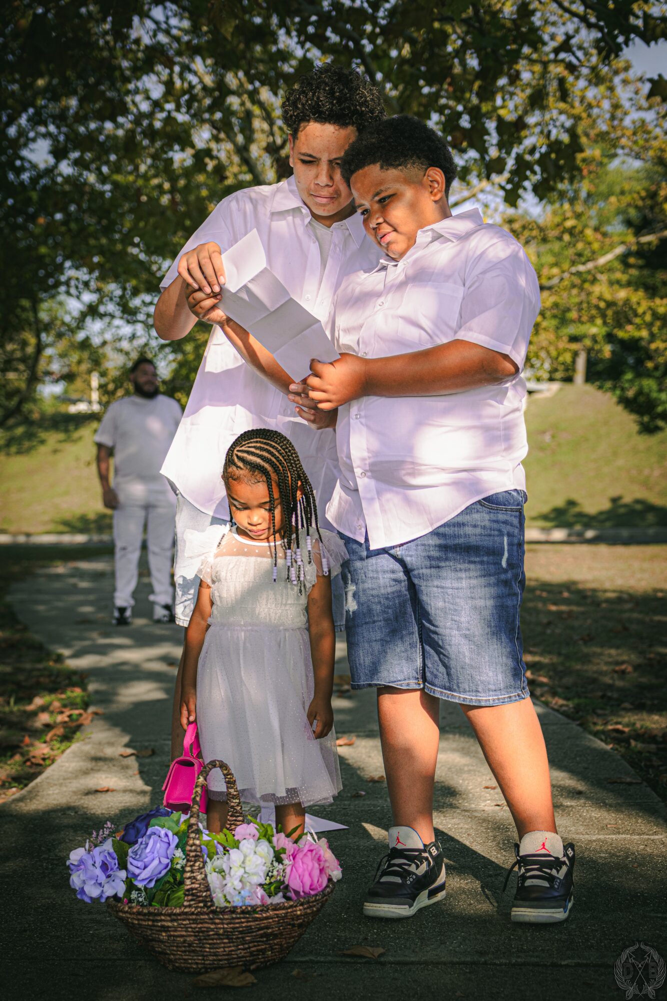 Two boys and a girl stand outdoors with a basket of flowers in front. The boys hold a photo, and the girl looks down.