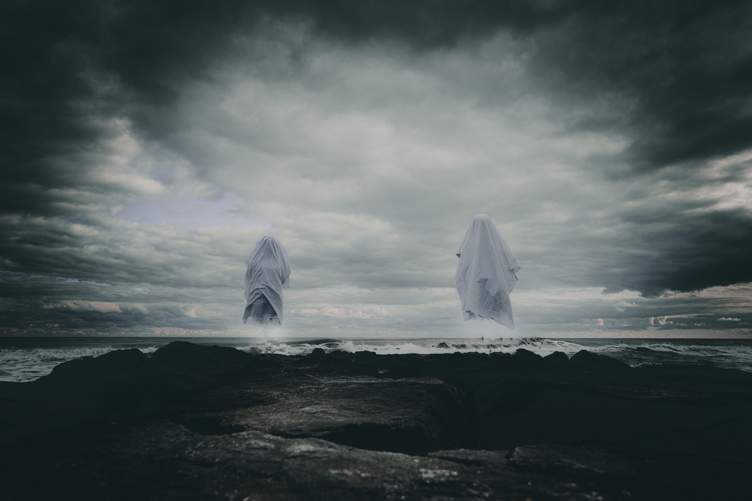 Two ghostly figures draped in white cloth stand on a dark shoreline under a cloudy sky.