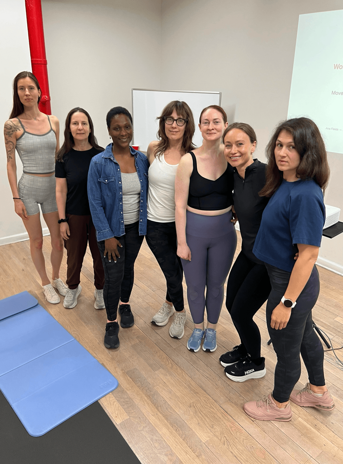 Seven women standing together in a room, some smiling, with a whiteboard and a projection screen in the background.