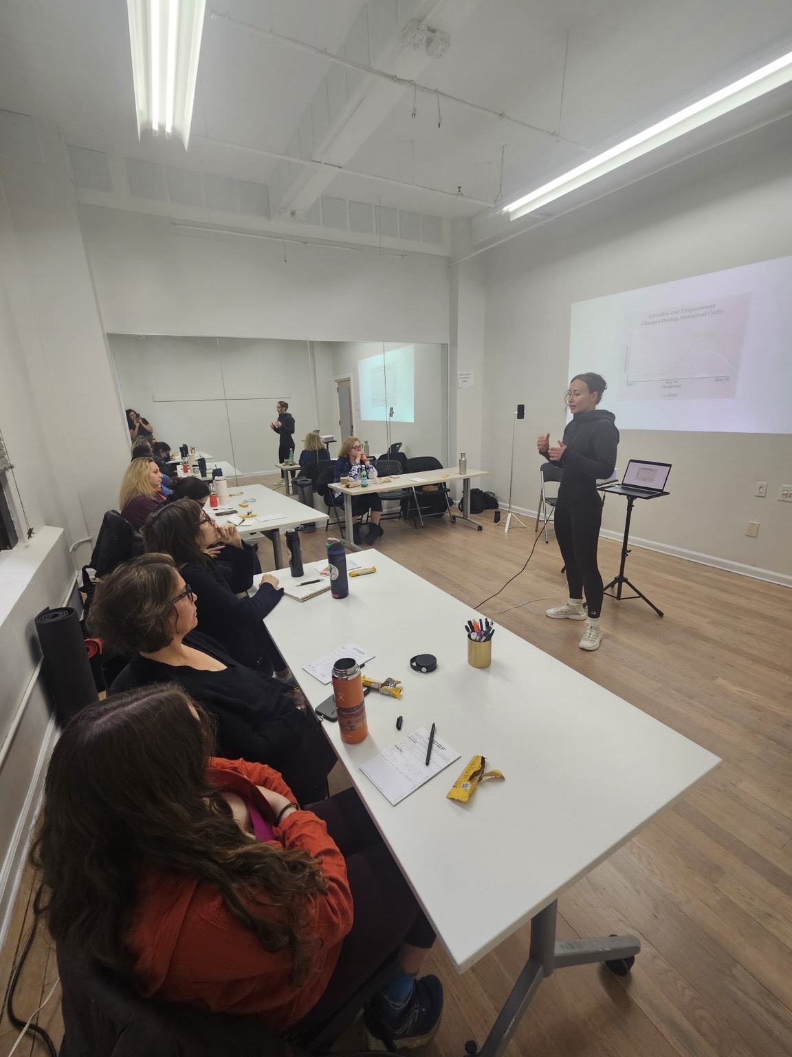 Person standing and presenting to seated group in a bright room with white walls and a projection screen.