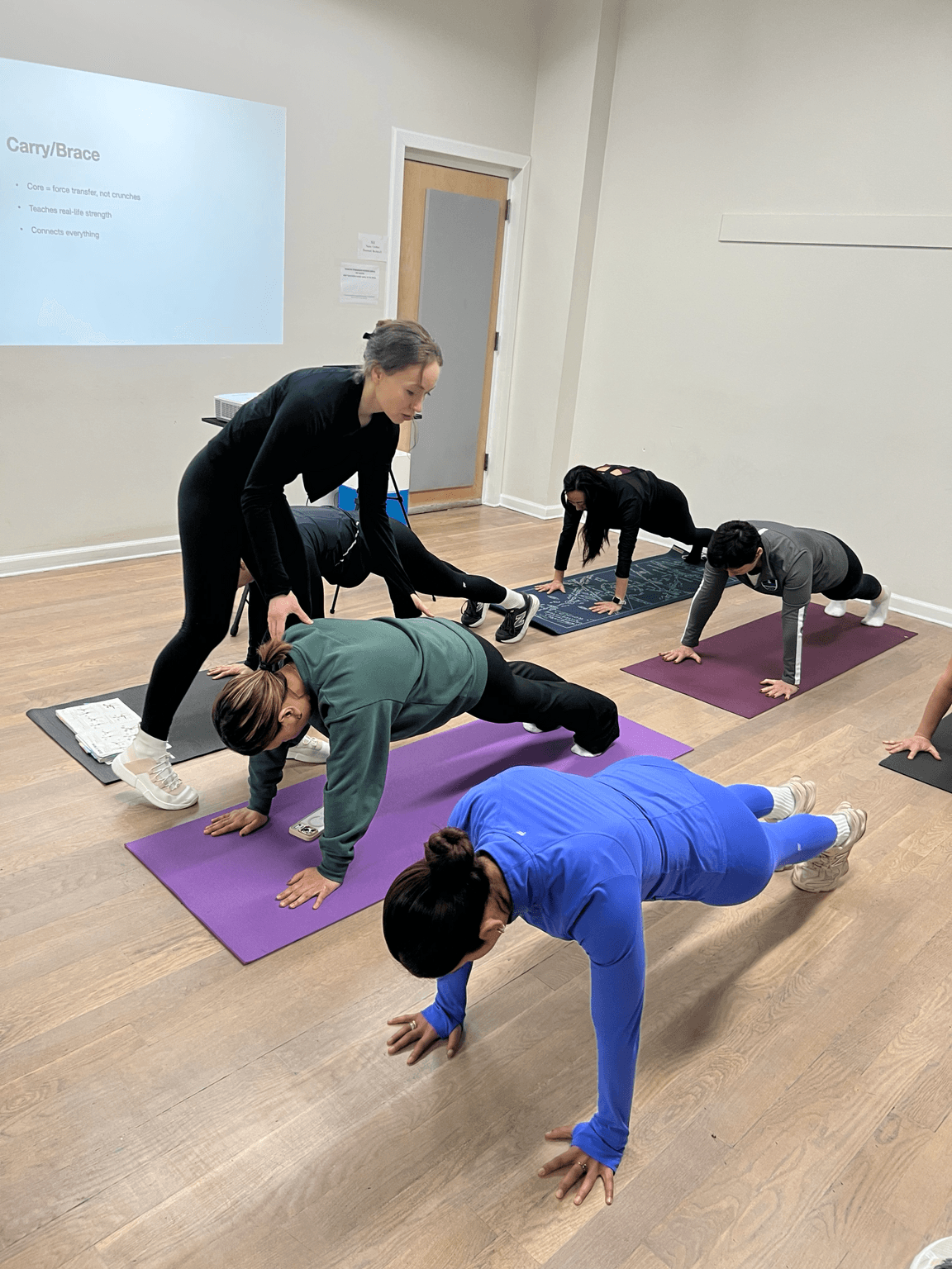 Group of people doing yoga in a room with a white wall and wooden floor, some on purple mats, instructor assisting.
