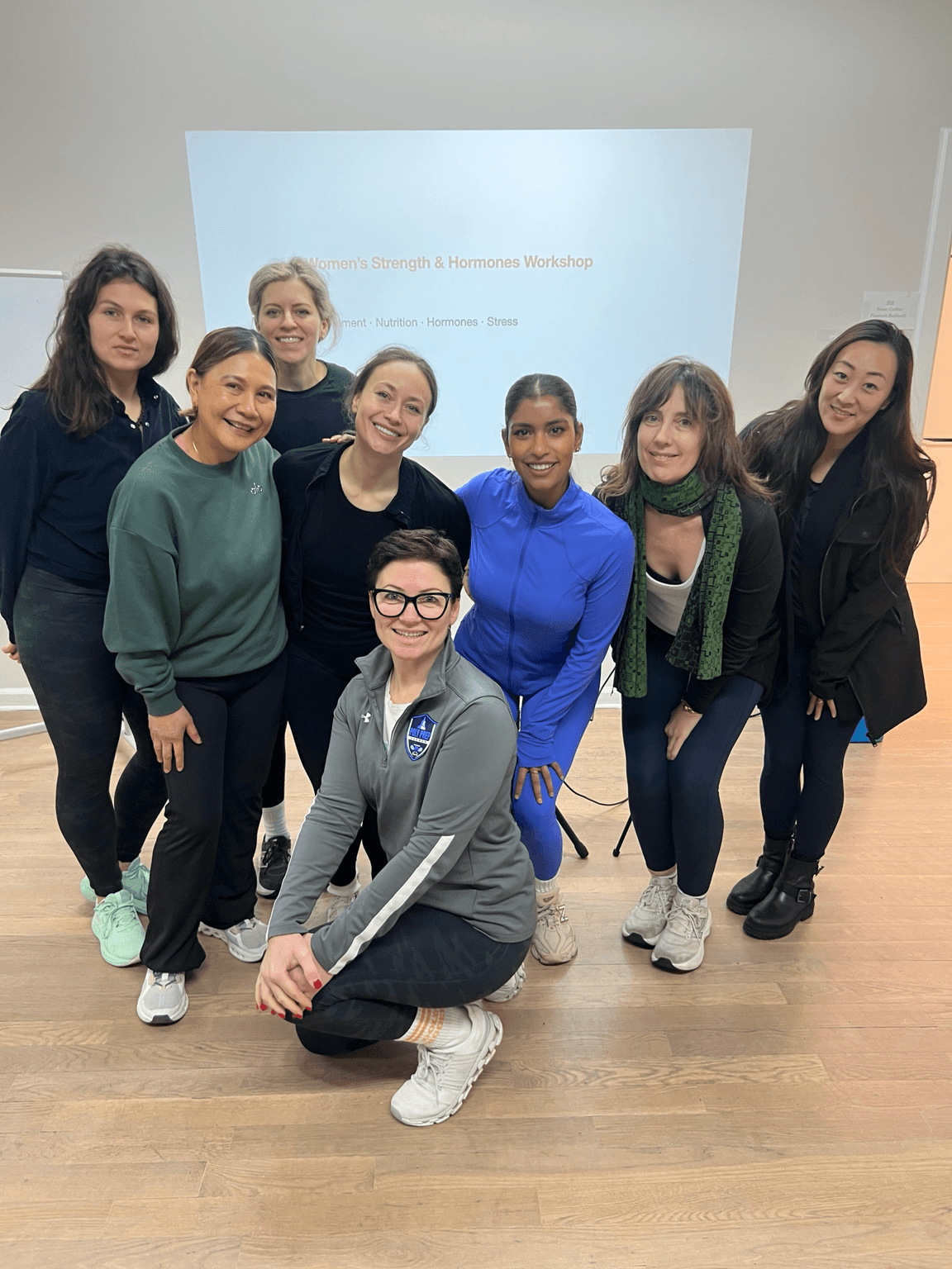 Group of nine women standing and sitting in a room with a presentation screen behind them.