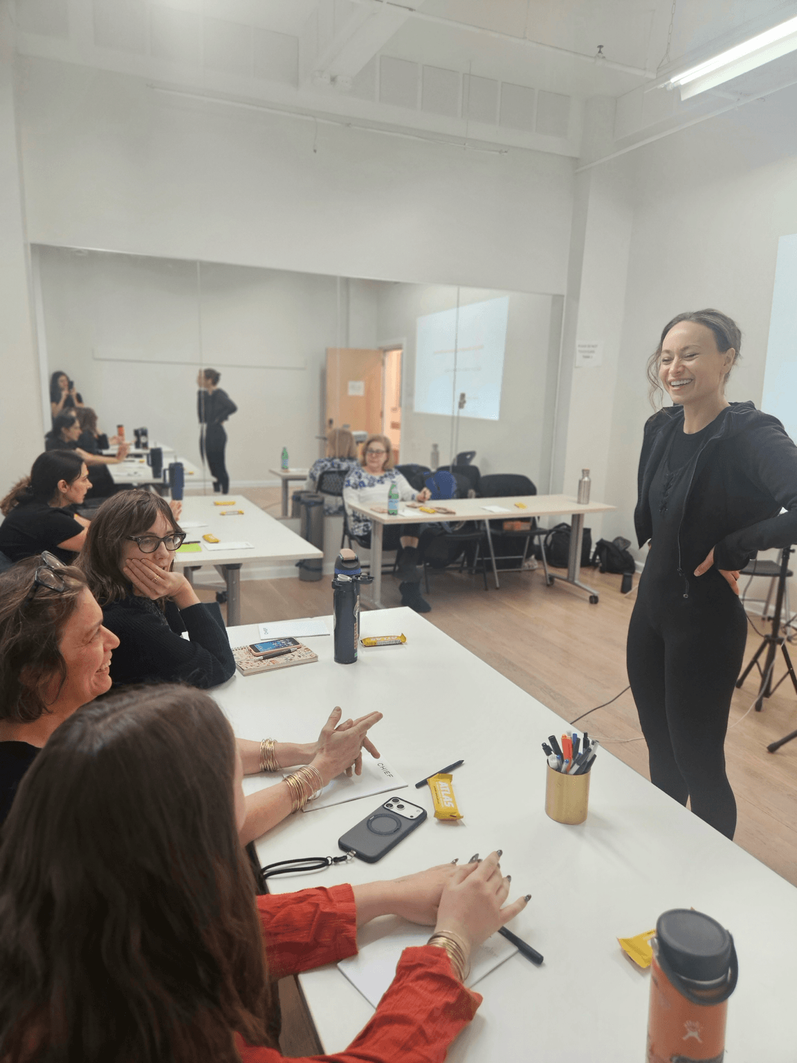 A woman stands at the front of a classroom, smiling, with students seated at tables listening and taking notes.