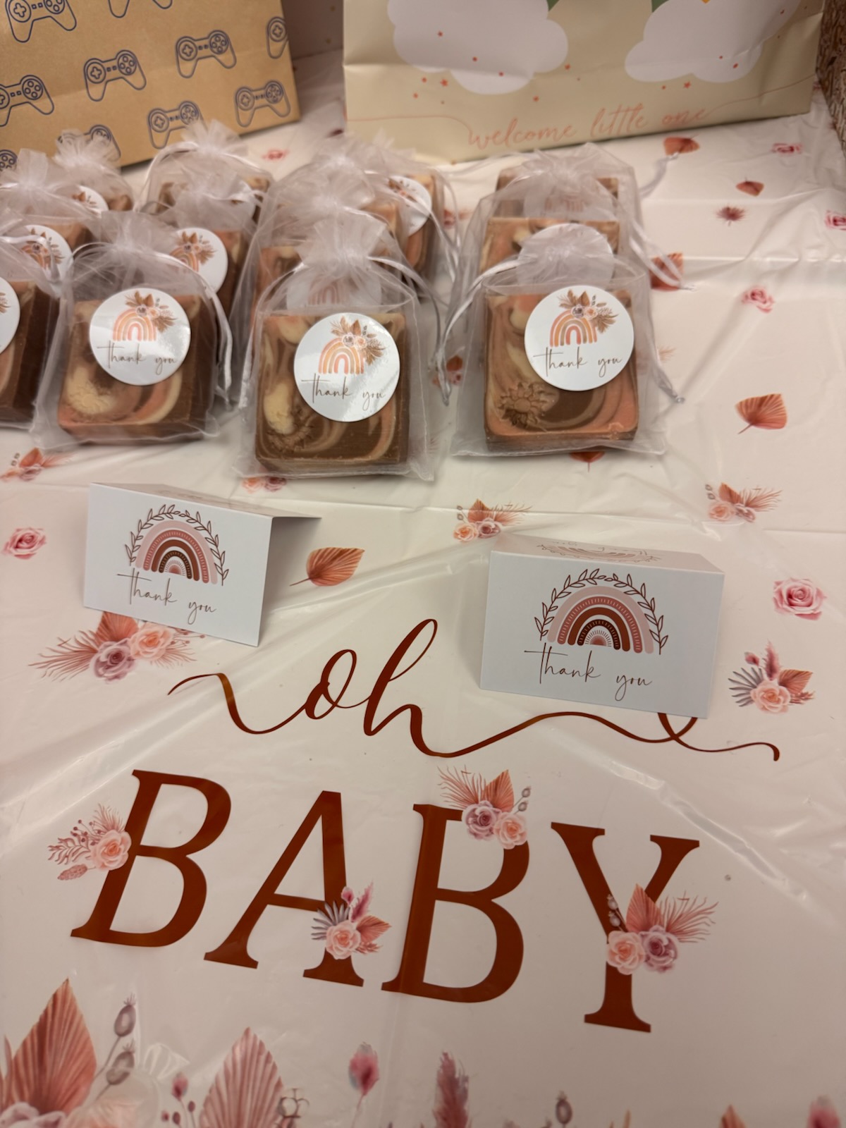 Cookies in clear bags with a tree logo, displayed on a table with a pink floral tablecloth and a sign reading 'oh BABY'.