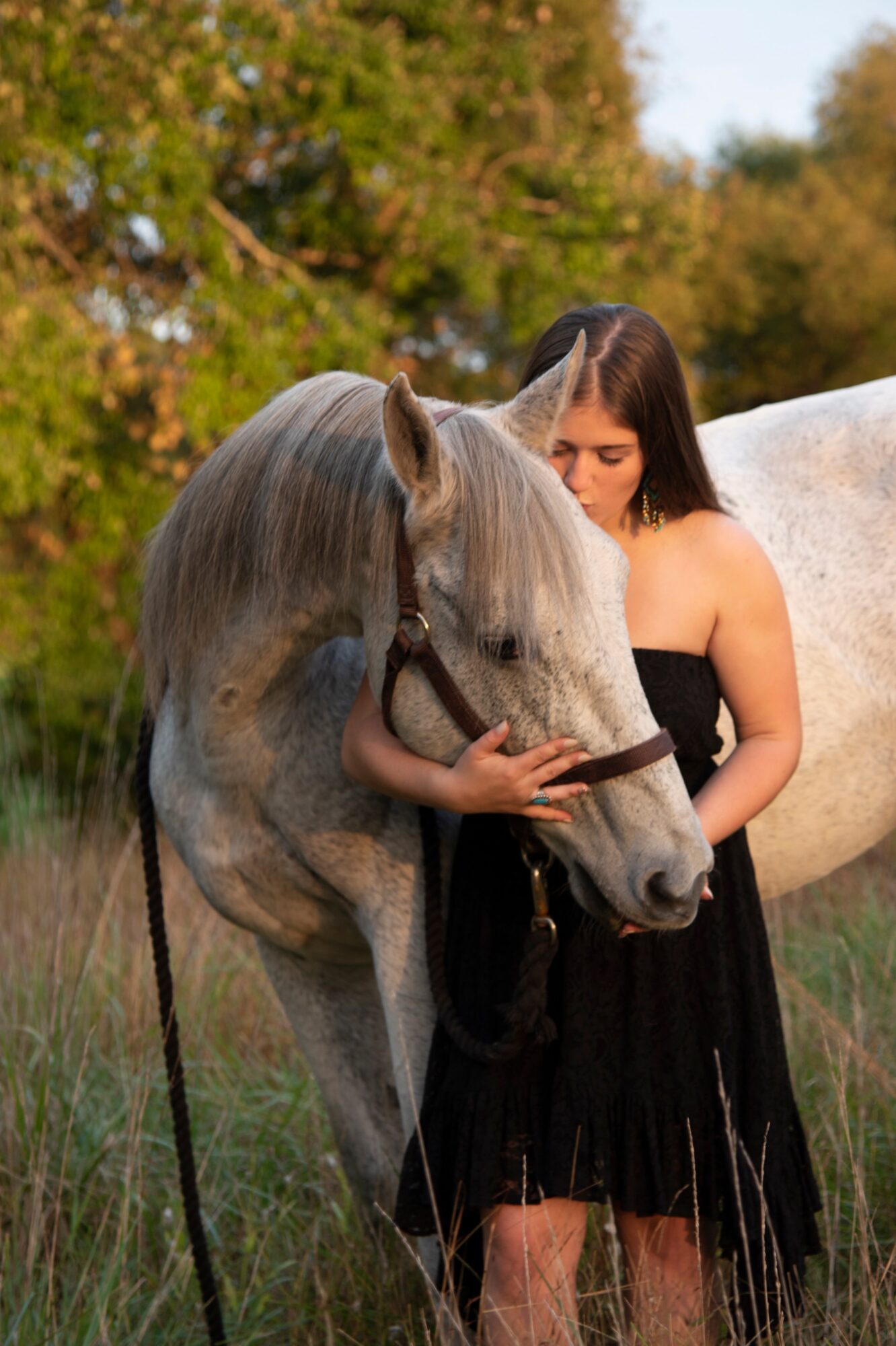 Young woman in black dress standing next to a gray horse outdoors, trees in background.