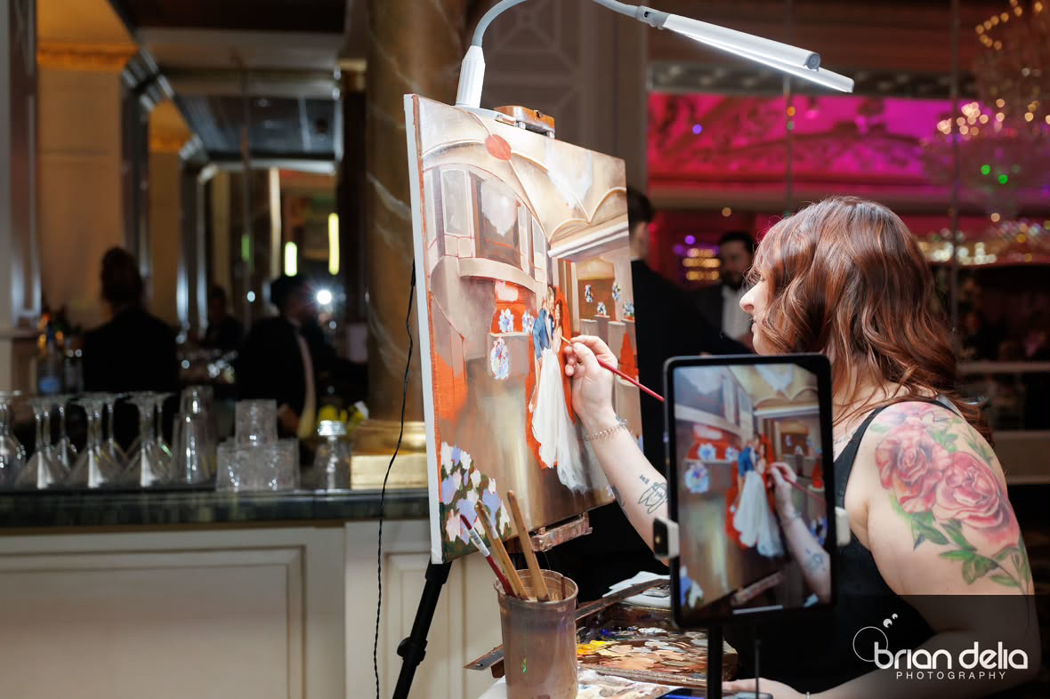 Person painting on canvas in a lively indoor setting with bar and people in background, holding a phone to record.