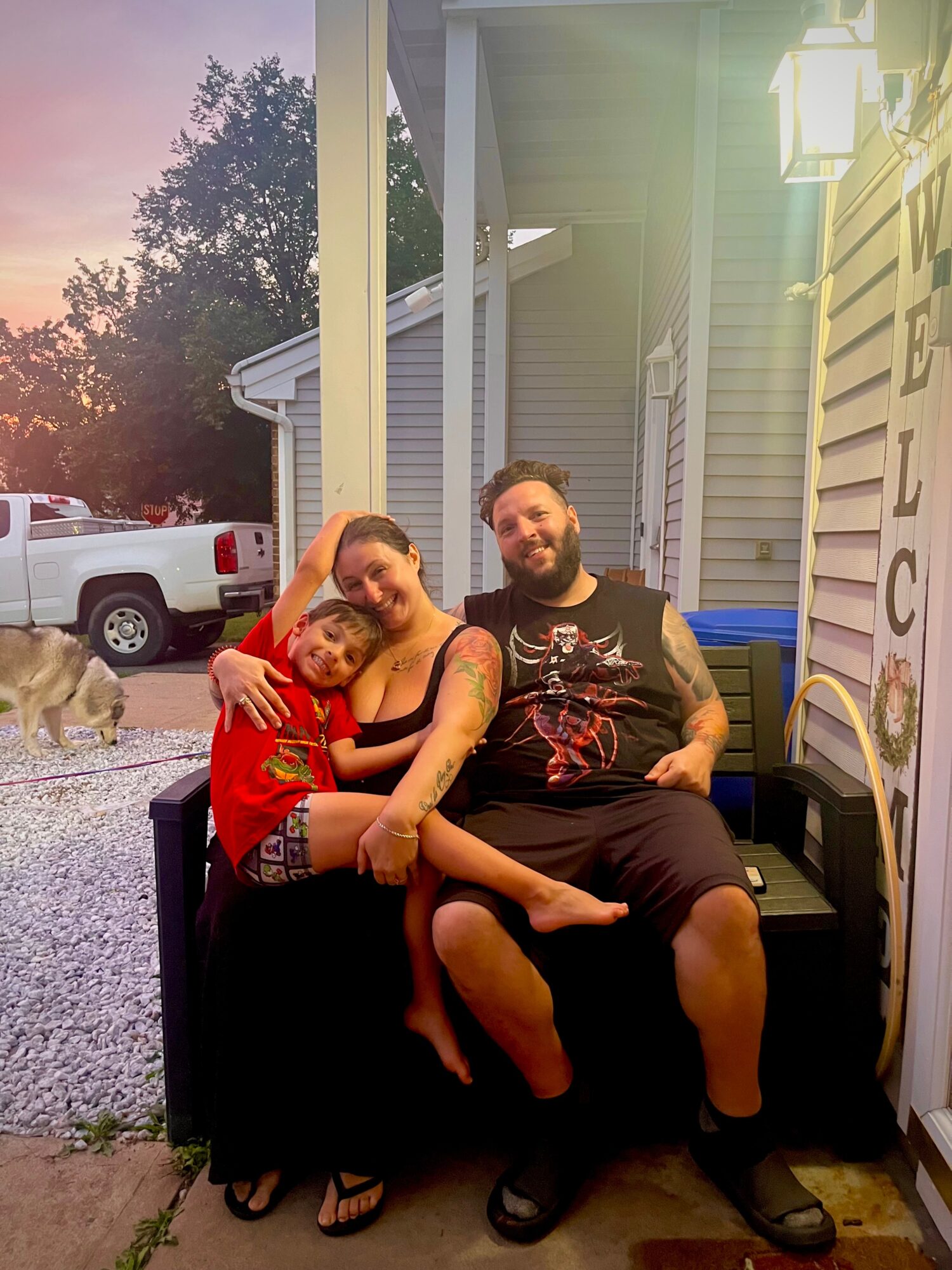 Two people sitting on a porch swing, smiling, with a child hugging them, during sunset outside a house.