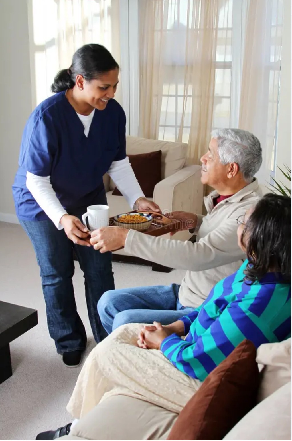 A woman in a blue uniform hands a tray with a cake and a mug to an elderly man sitting on a sofa, with a woman in a striped sweater nearby.