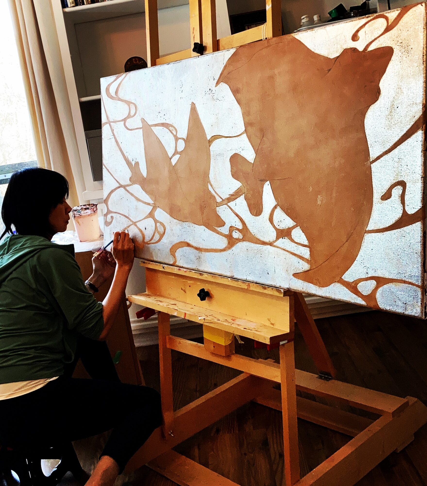 Person painting a large canvas with animal silhouettes, sitting indoors near a window, using a brush, with a wooden easel.