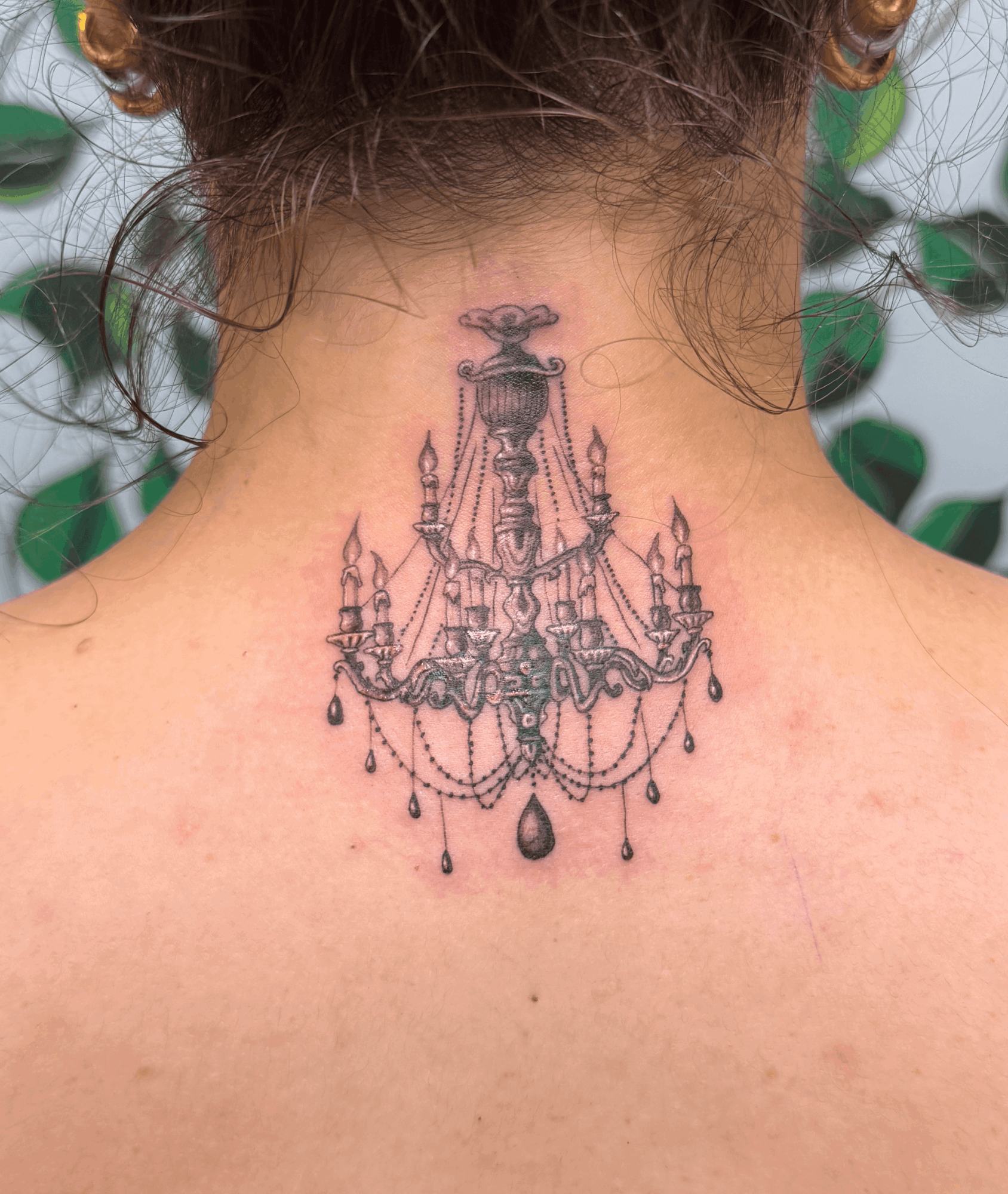 Back of a person with a chandelier tattoo on the upper back, detailed with hanging crystals and ornate design.