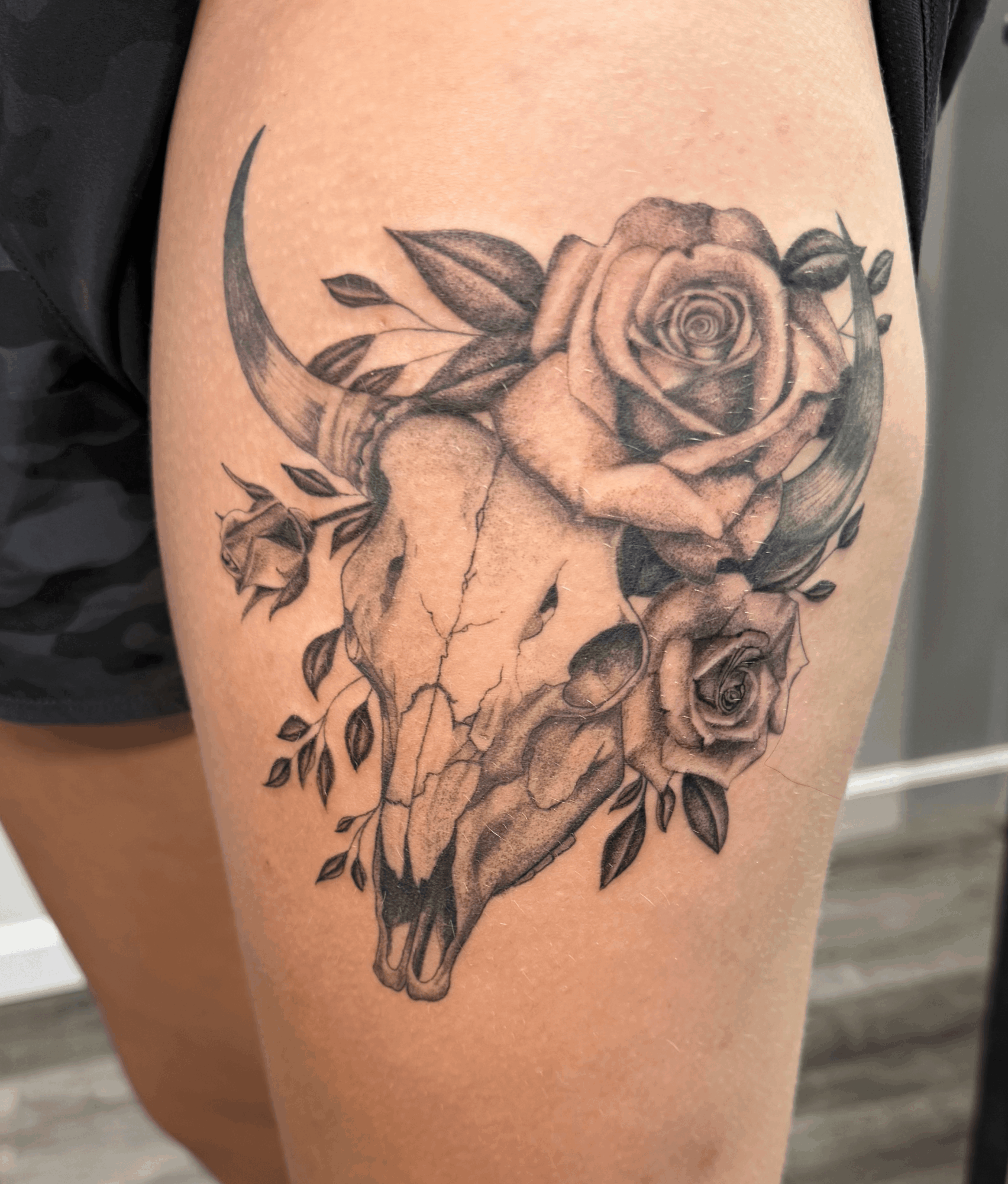 Tattoo of a skull with roses and leaves on thigh, detailed black and gray shading, crescent moon in background.