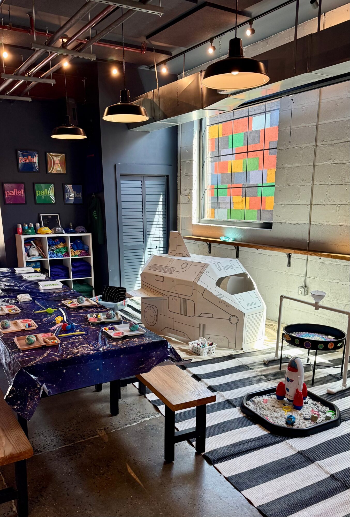 Indoor space with tables, artwork, and a large window with colorful grid display, black and white striped rug, and laundry machines.