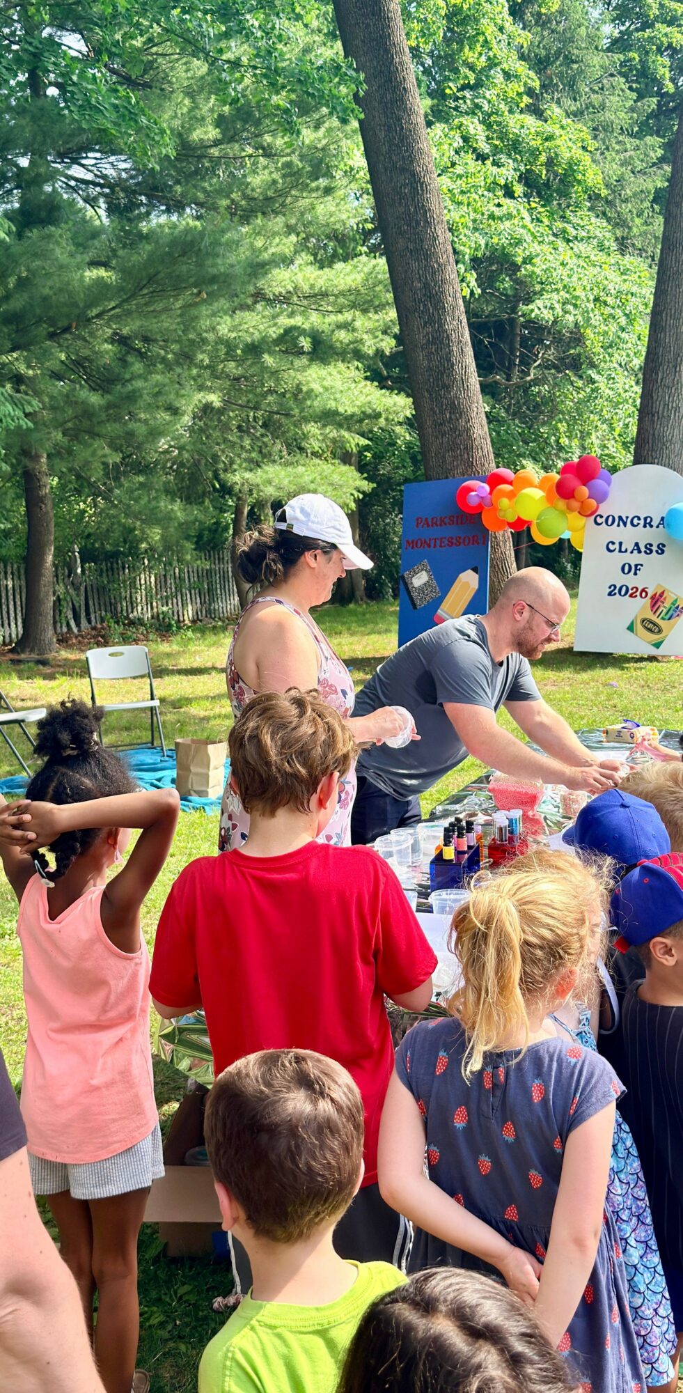 Children gather outdoors near a table with balloons, trees in background, and a colorful sign, during daytime.
