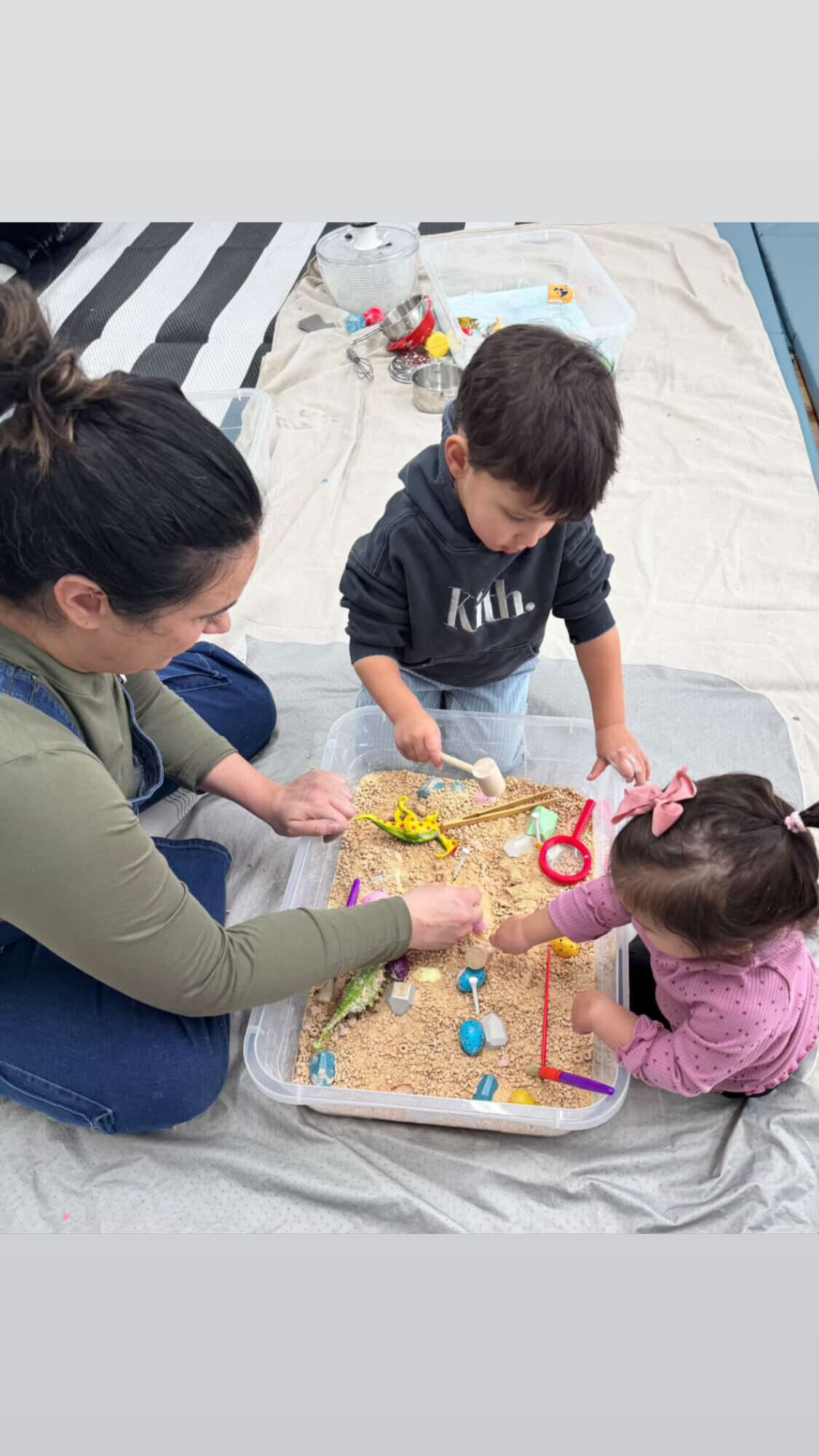 Two children and an adult playing a board game on a blanket outdoors, with toys and snacks nearby.