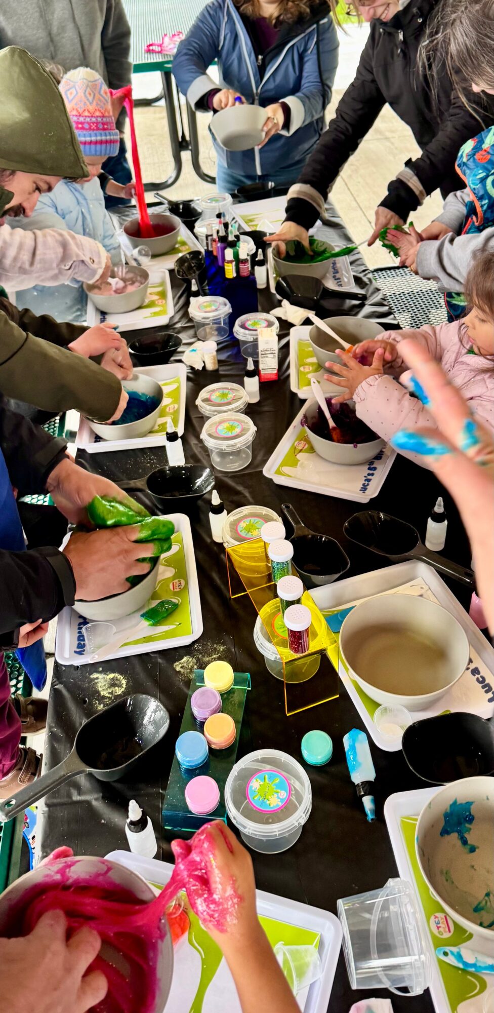 Children and adults gather around a table with art supplies, painting and decorating objects, with various bottles and containers present.