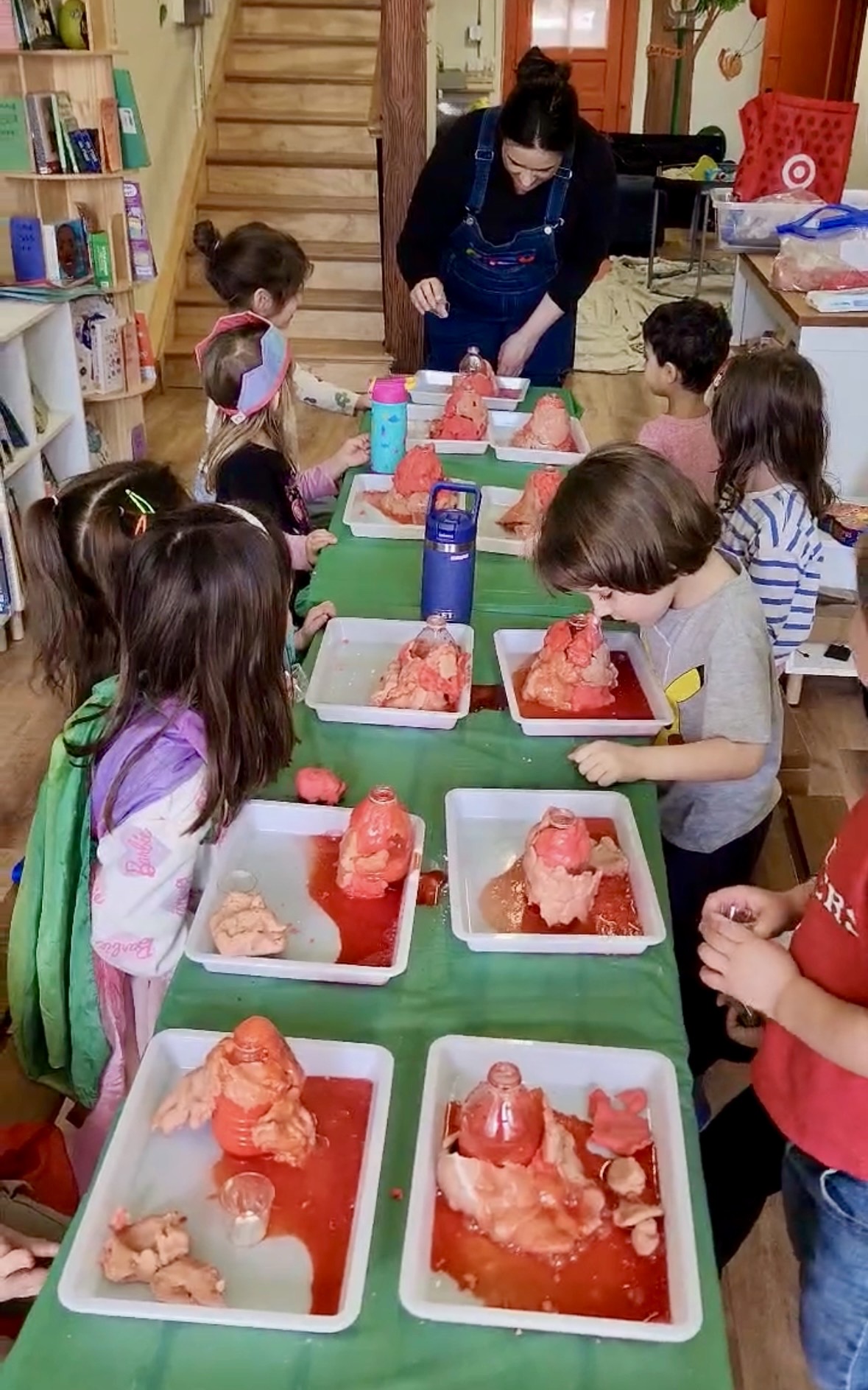Children sit at a table with a woman, making ice cream sundaes with toppings, in a colorful room with bookshelves.