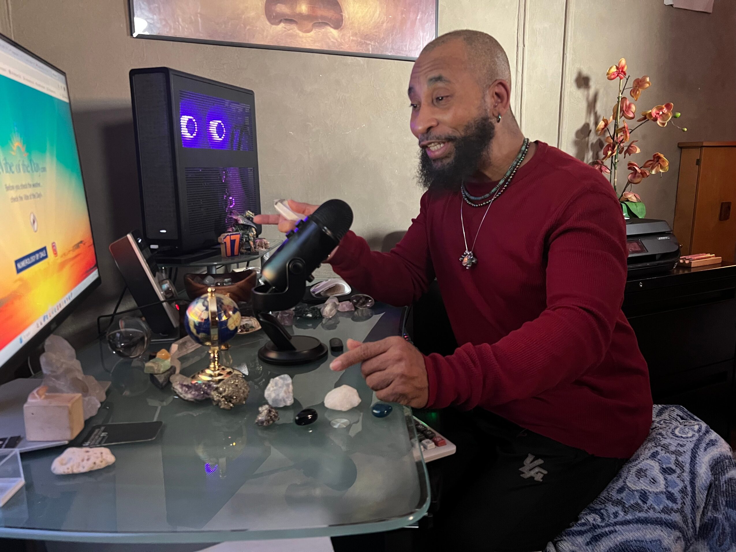 Man with a beard and red sweater sitting at a desk with a microphone, computer, and decorative items, smiling.