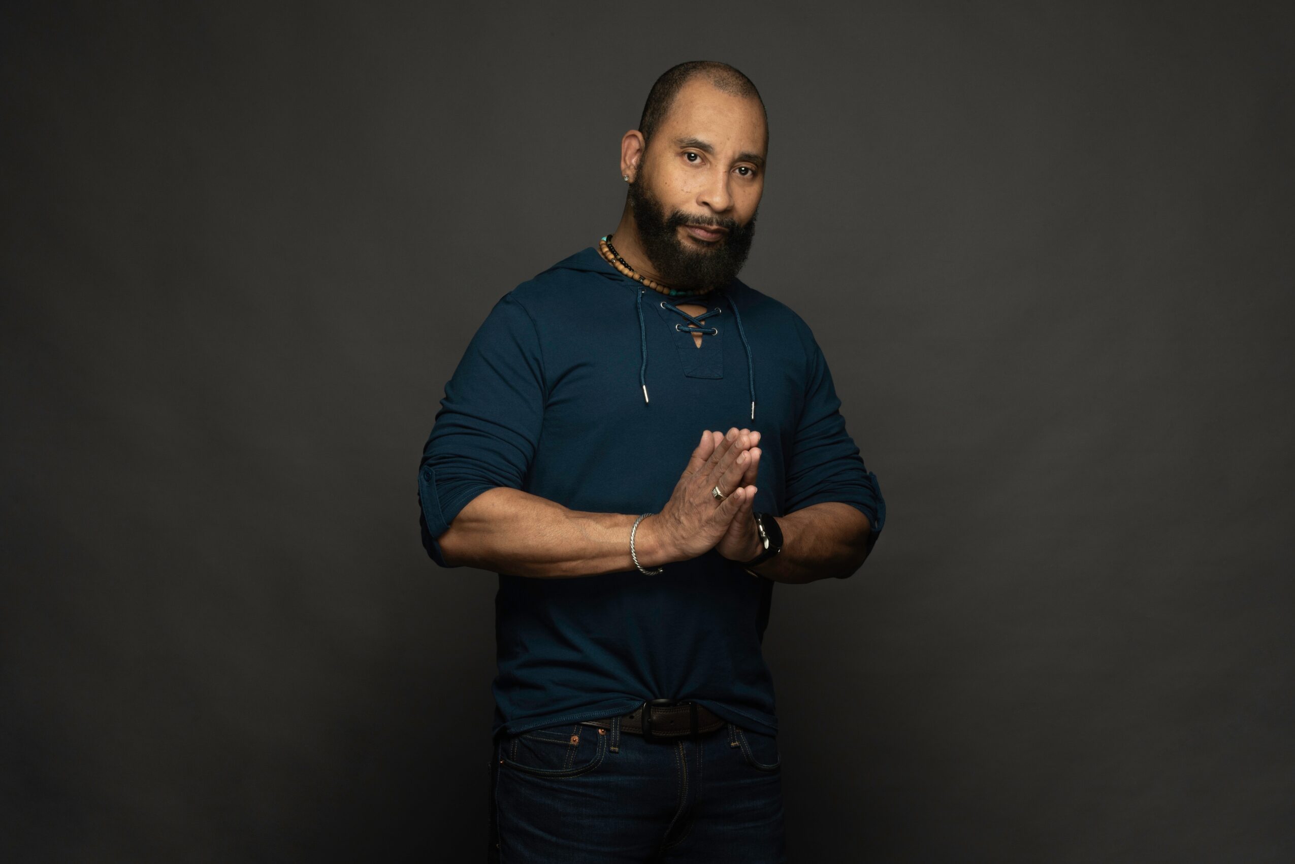 Man with a beard and mustache, wearing a dark blue shirt, standing with hands pressed together, against a dark background.