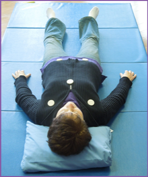 Person lying on a blue mat with head on a pillow, arms outstretched, wearing a dark sweater and light pants.