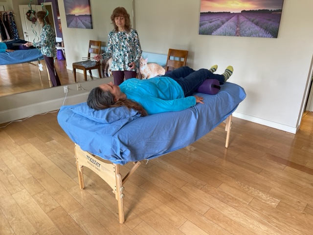 Person lying face down on a massage table with a blue cover, in a room with wooden floor and wall art.